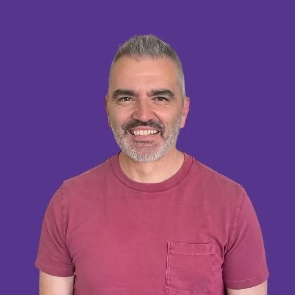 A smiling man with gray hair and beard wearing a maroon T-shirt against a purple background.