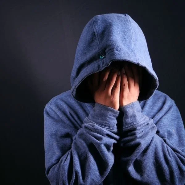 Person wearing a blue hoodie with the hood up, covering their face with their hands, against a dark background.