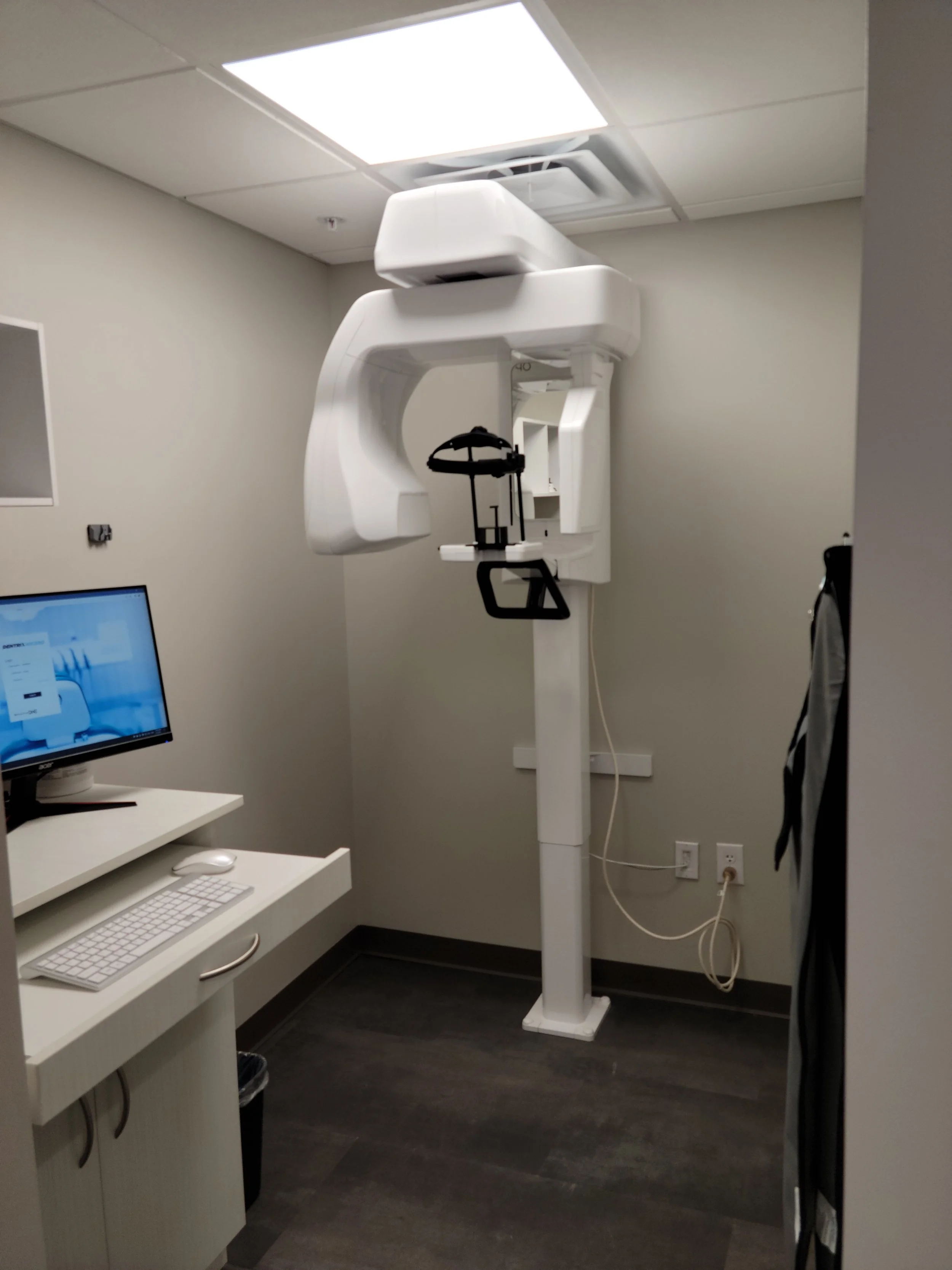 Dental X-ray machine in an examination room with a computer and keyboard on a desk.