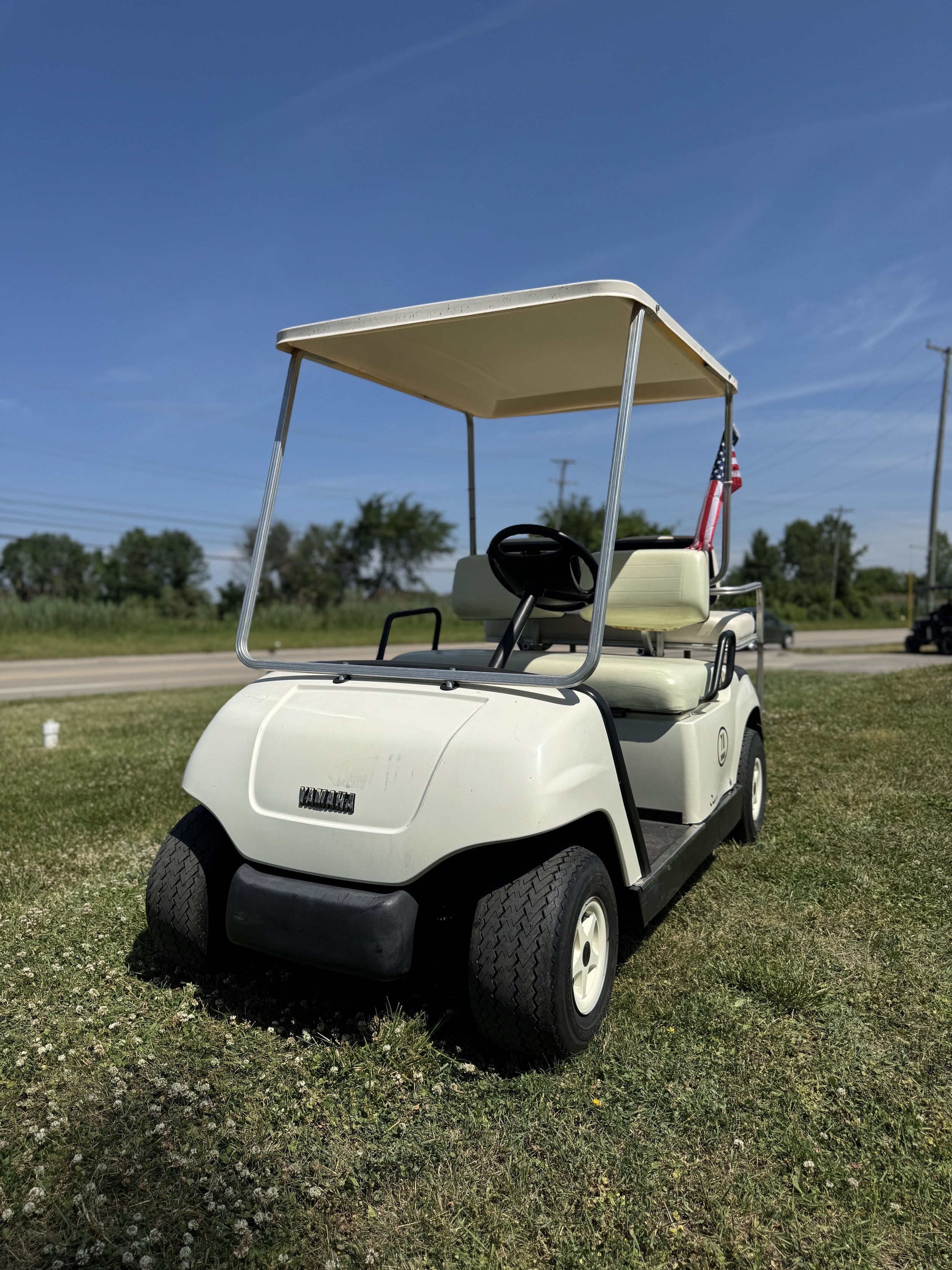 White golf cart parked on grass under a blue sky with trees and utility poles in the background.