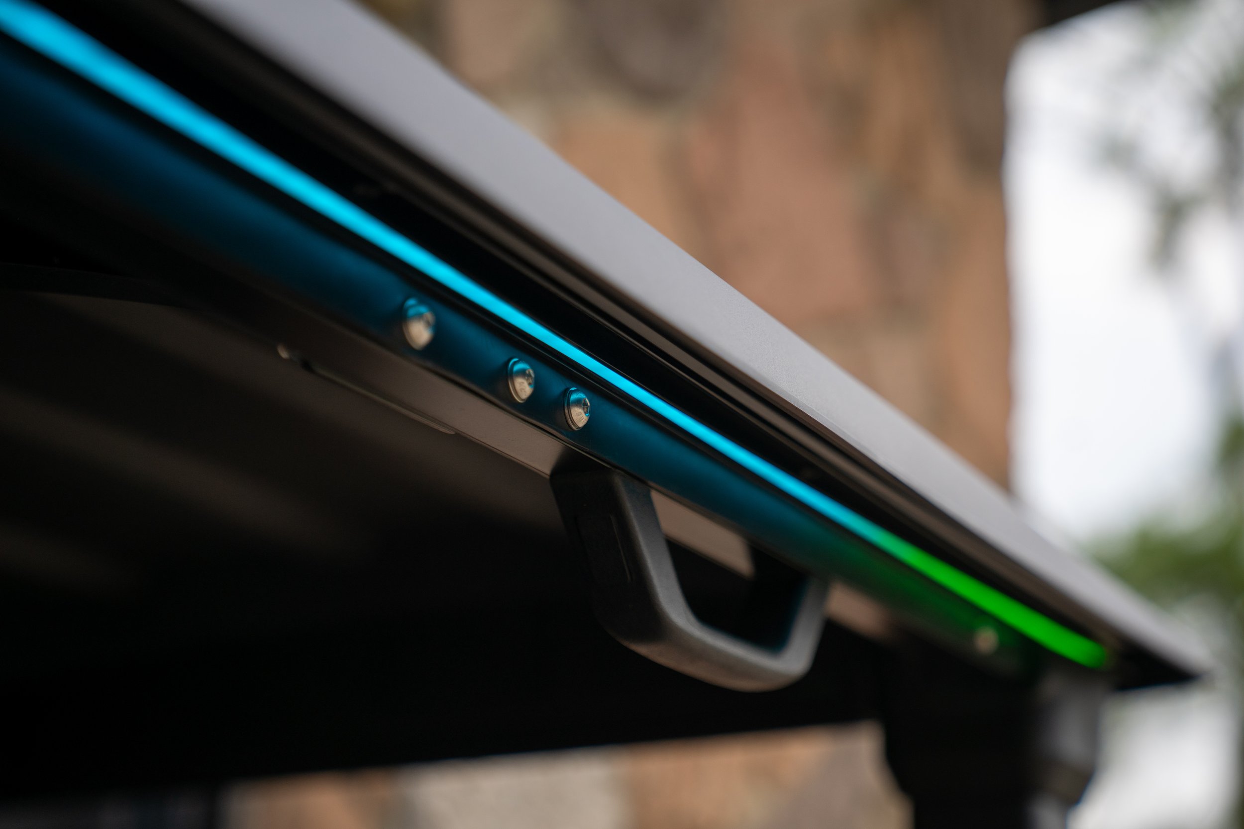 Close-up of a modern electric skateboard with blue and green LED lights along the underside, showing a black handle and metal components.
