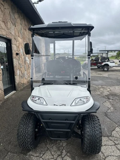Front view of a white golf cart with a clear windshield, black roof, and large tires parked outside a stone building and other vehicles in the background.