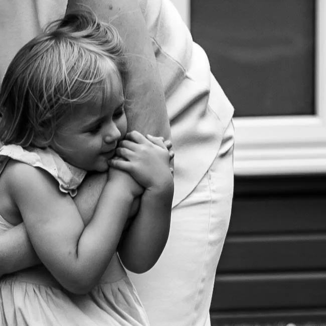 A young girl hugging an adult's arm, resting her head against it, with a window in the background.
