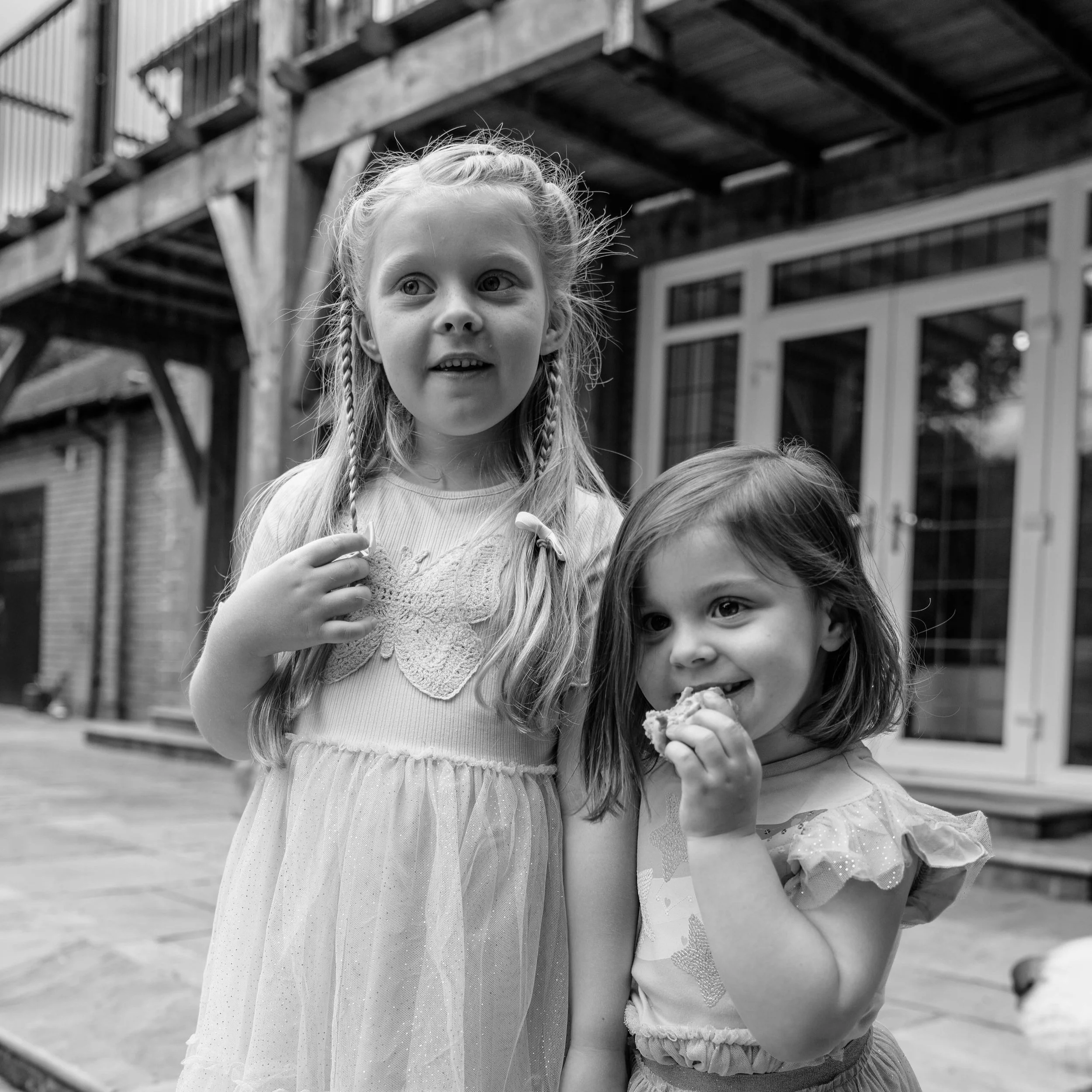 Two young girls standing outside, one with long braided hair and the other with short hair, both holding snacks. They are near a building with large windows and a wooden deck.