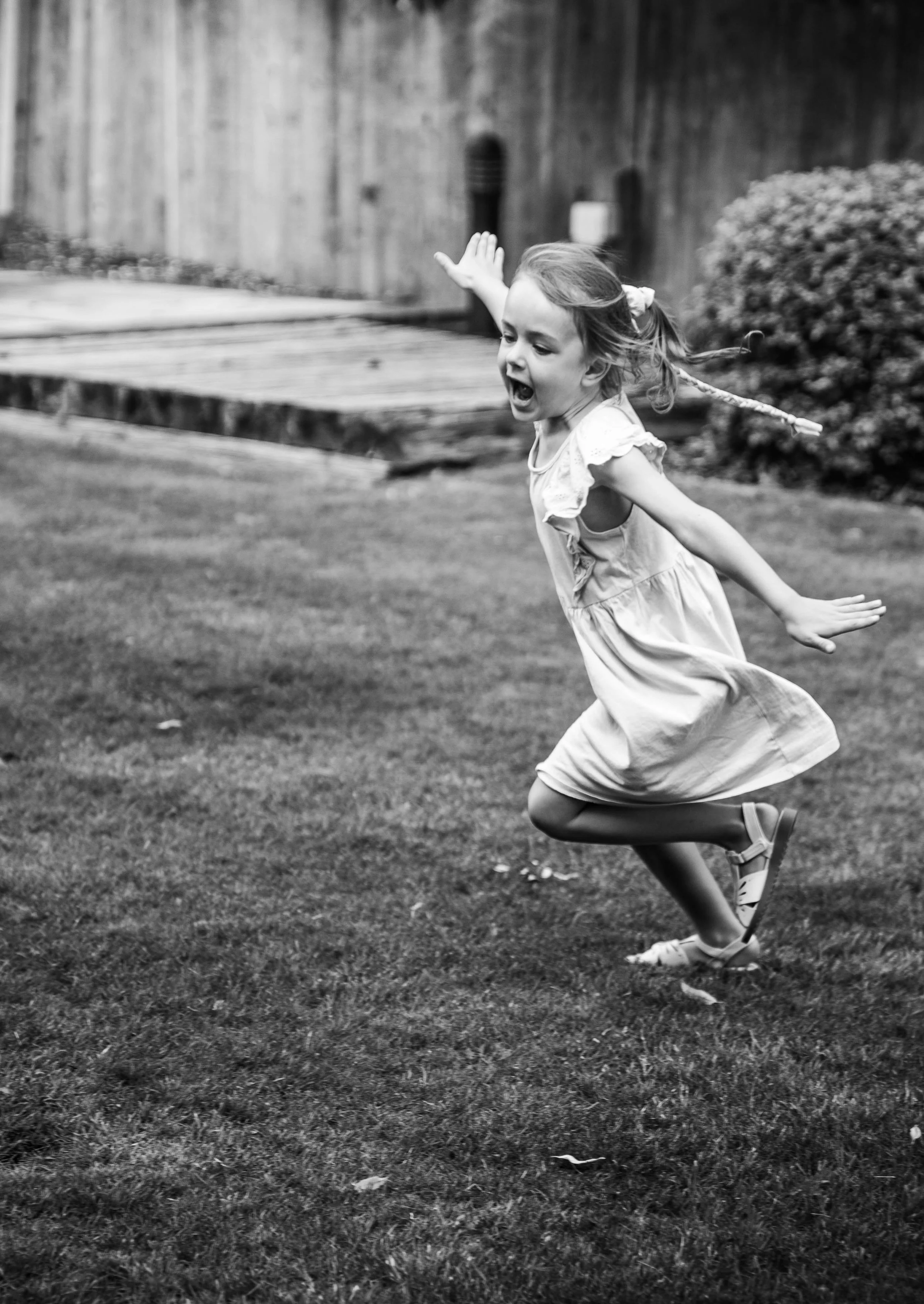 A young girl with braided hair is playing and running on the grass in a backyard, with a wooden fence and bush in the background. She appears to be excited or happy.