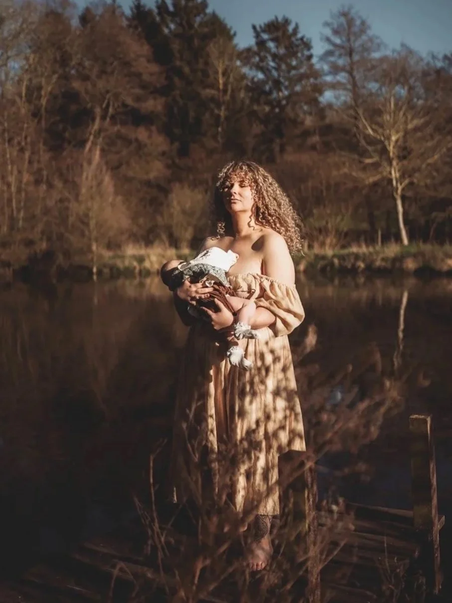 A woman with curly hair wearing a peach-colored dress stands in front of a body of water, holding a baby wrapped in a blanket. The scene is outdoors with trees in the background and the lighting suggests it is late afternoon or early evening.