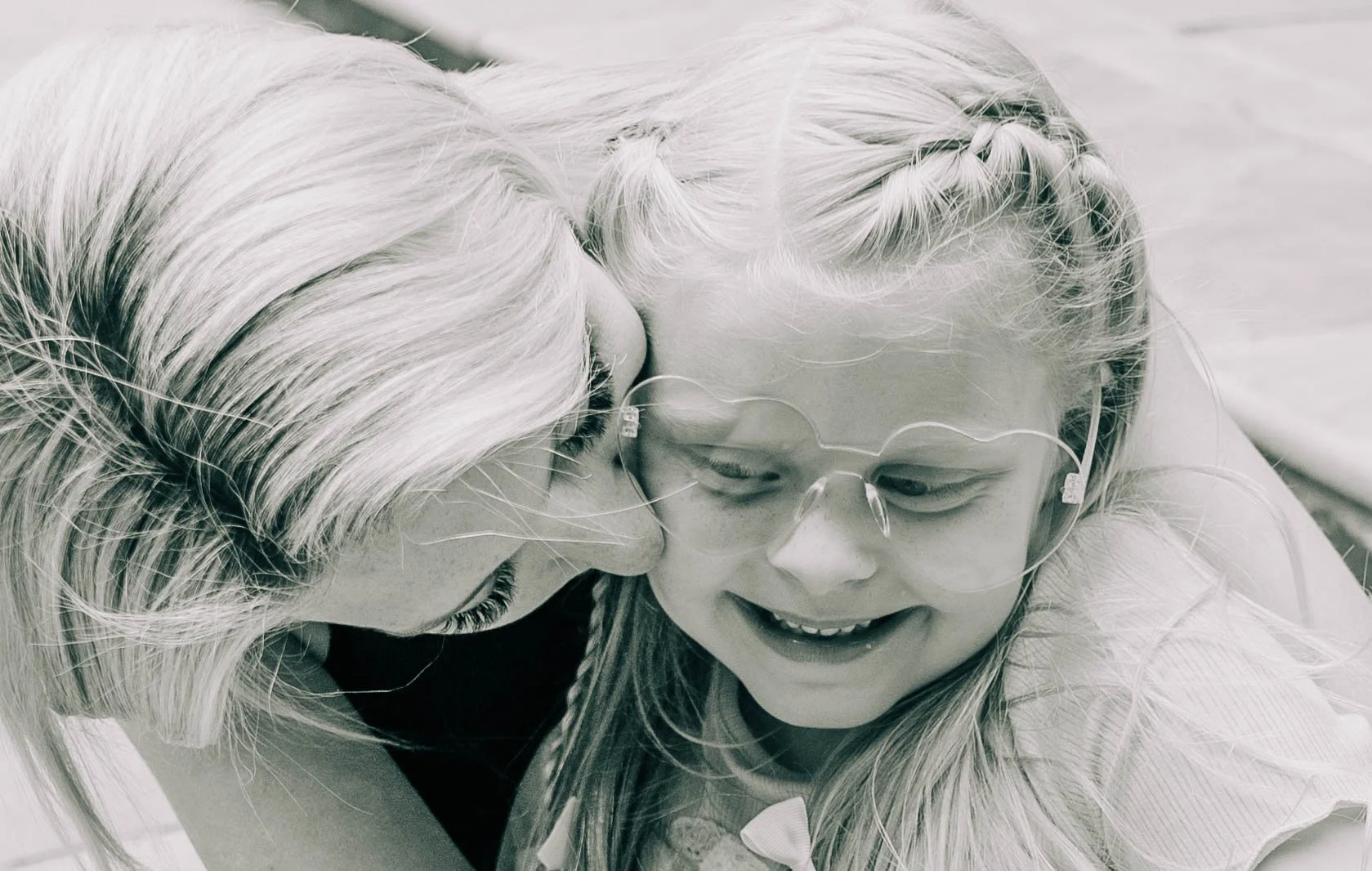 A woman and young girl are hugging each other, smiling and sharing an affectionate moment. The woman has blond hair and the girl has blond hair with a braid, and they are both wearing glasses.