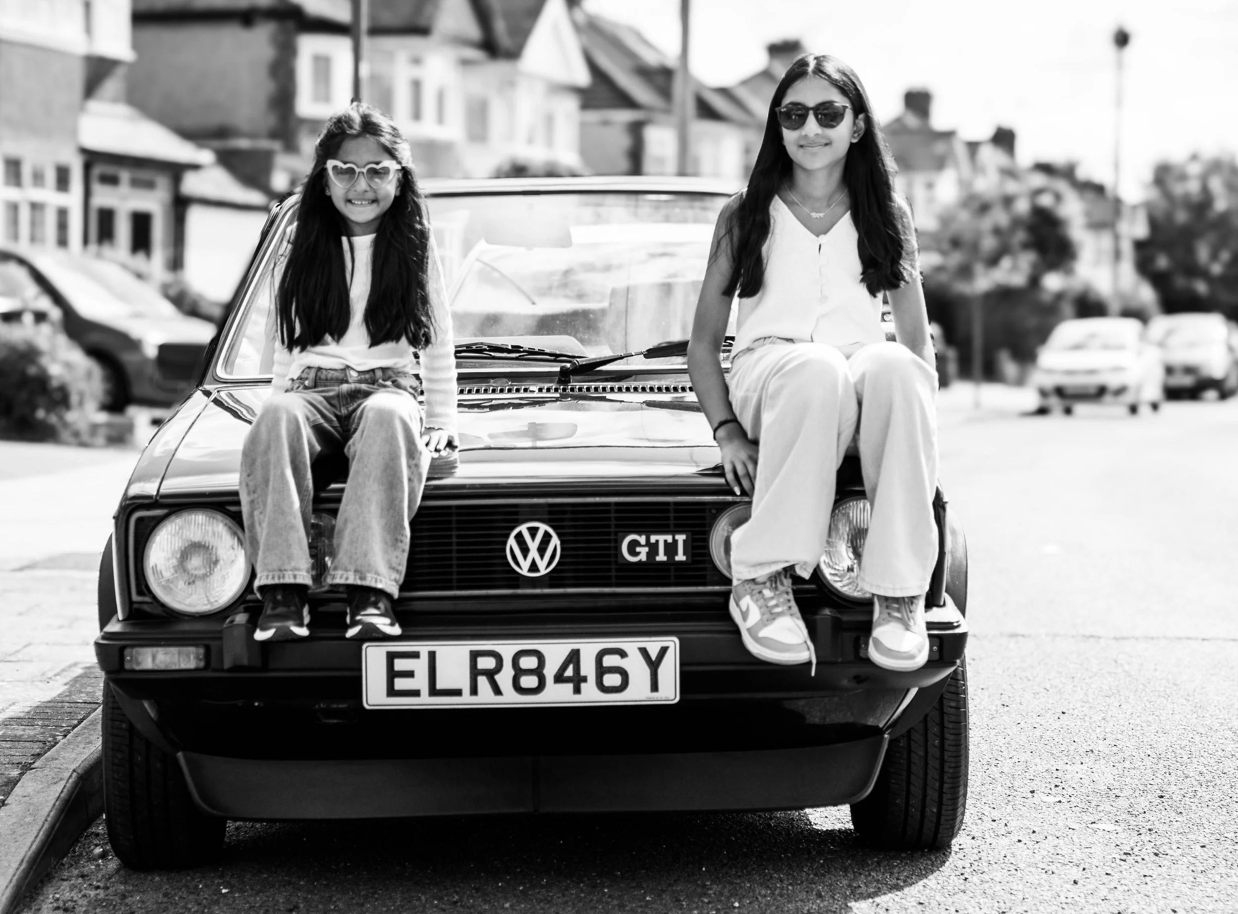 Two young girls wearing sunglasses sitting on the hood of a vintage Volkswagen GTI car parked on a residential street, smiling at the camera.