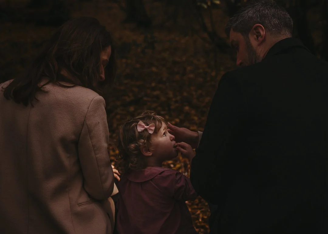 A family of three, a woman, man, and young girl, is gathered outdoors at night with autumn leaves on the ground. The man and woman are looking at the young girl, who is touching her lips, and they seem to be in a tender moment.