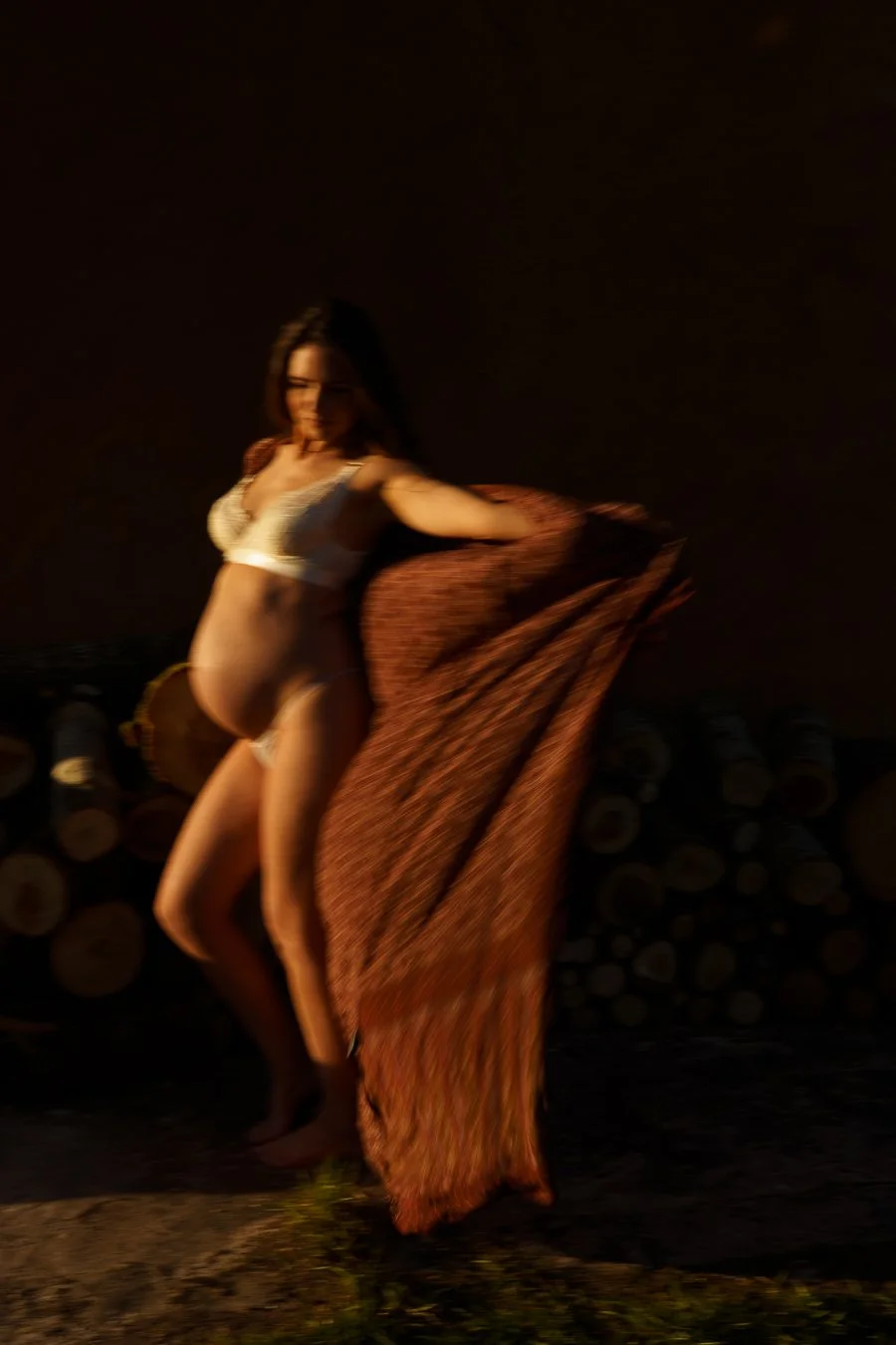 A pregnant woman in lingerie standing outdoors at night, holding a patterned fabric, with a background of stacked logs.