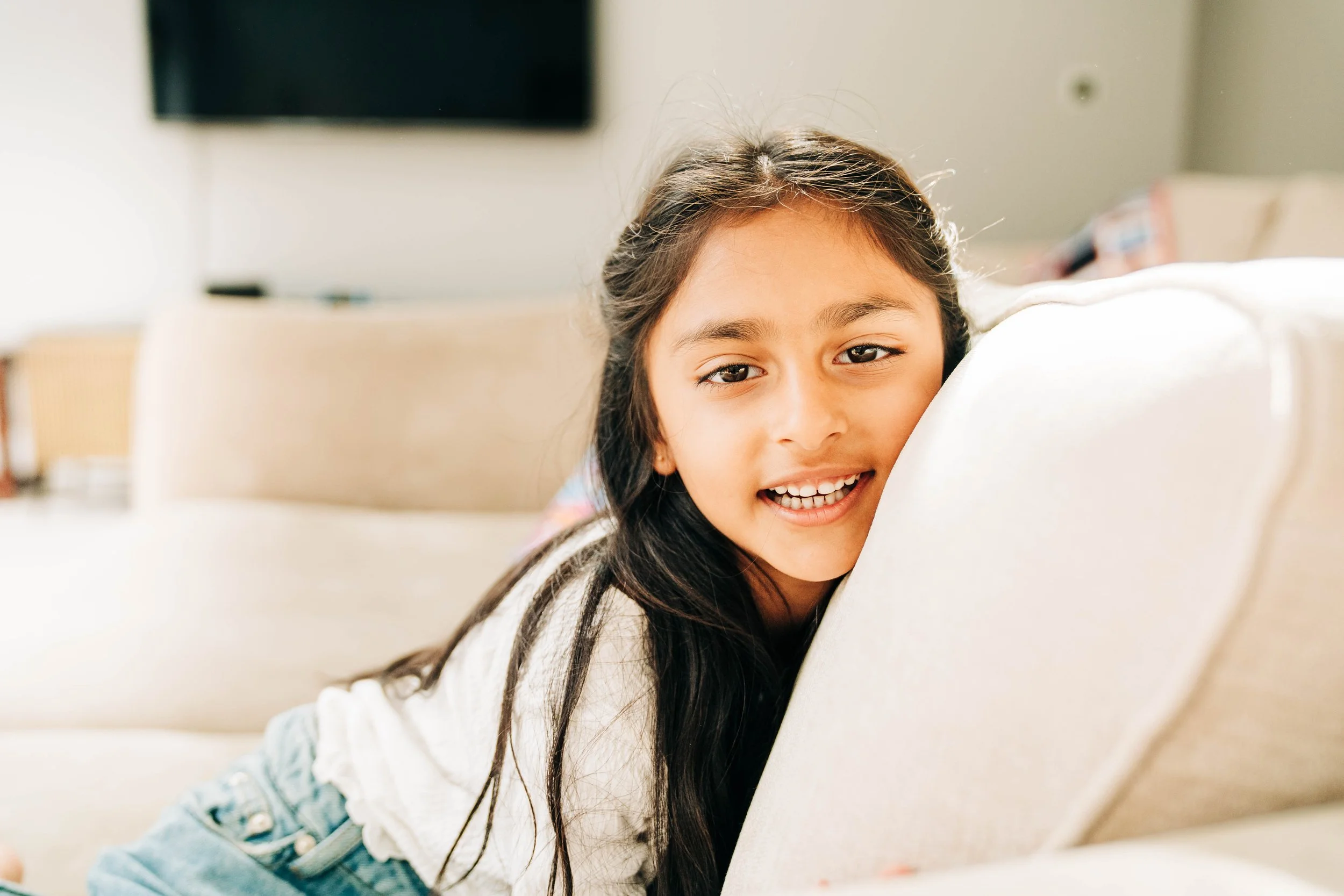 A young girl with long dark hair smiling and leaning on the back of a light-colored sofa in a living room.