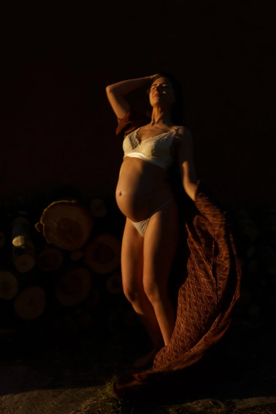 A woman lying on her side outdoors at sunset, wearing lingerie, with a stack of logs behind her.