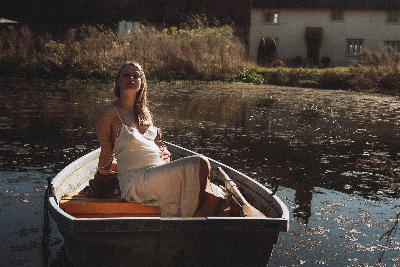 A woman wearing a light-colored dress sitting in a small boat on a calm river during late afternoon or early evening.