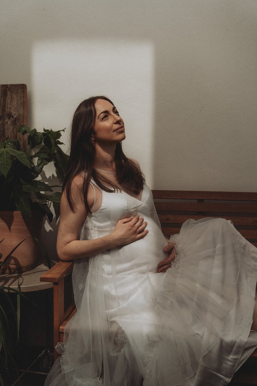 A pregnant woman in a white satin and tulle dress sitting on a wooden bench indoors, with a plant and wooden paneling nearby, looking thoughtfully to the side with a soft light illuminating her face.