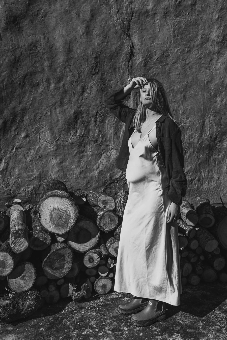 A woman in a long satin dress and a jacket standing outdoors against a textured rock wall, next to a pile of logs.