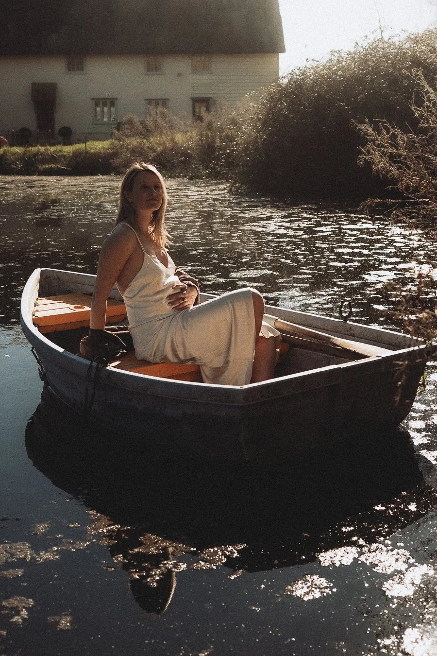 A woman in a white dress sitting in a small boat on a calm river during sunset.