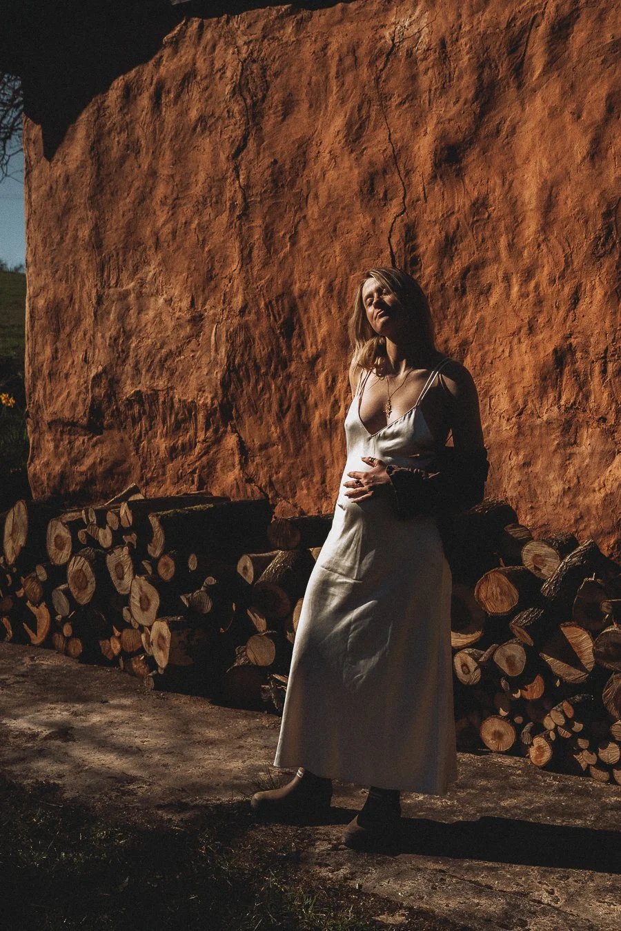A woman in a white dress standing outdoors against a large red rock wall with stacked cut logs nearby.