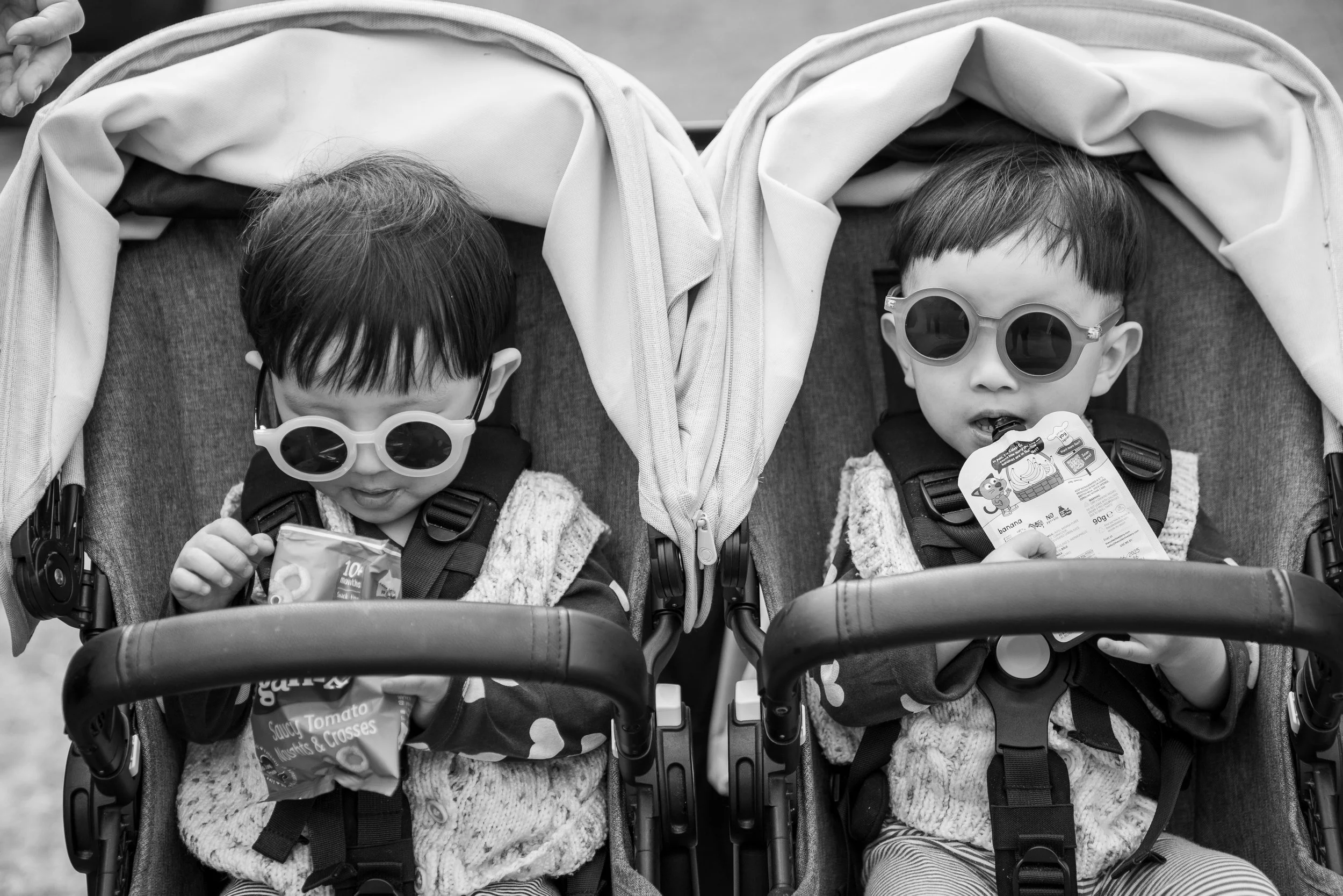 Two young children sitting in a double stroller, wearing sunglasses and holding snack packages.