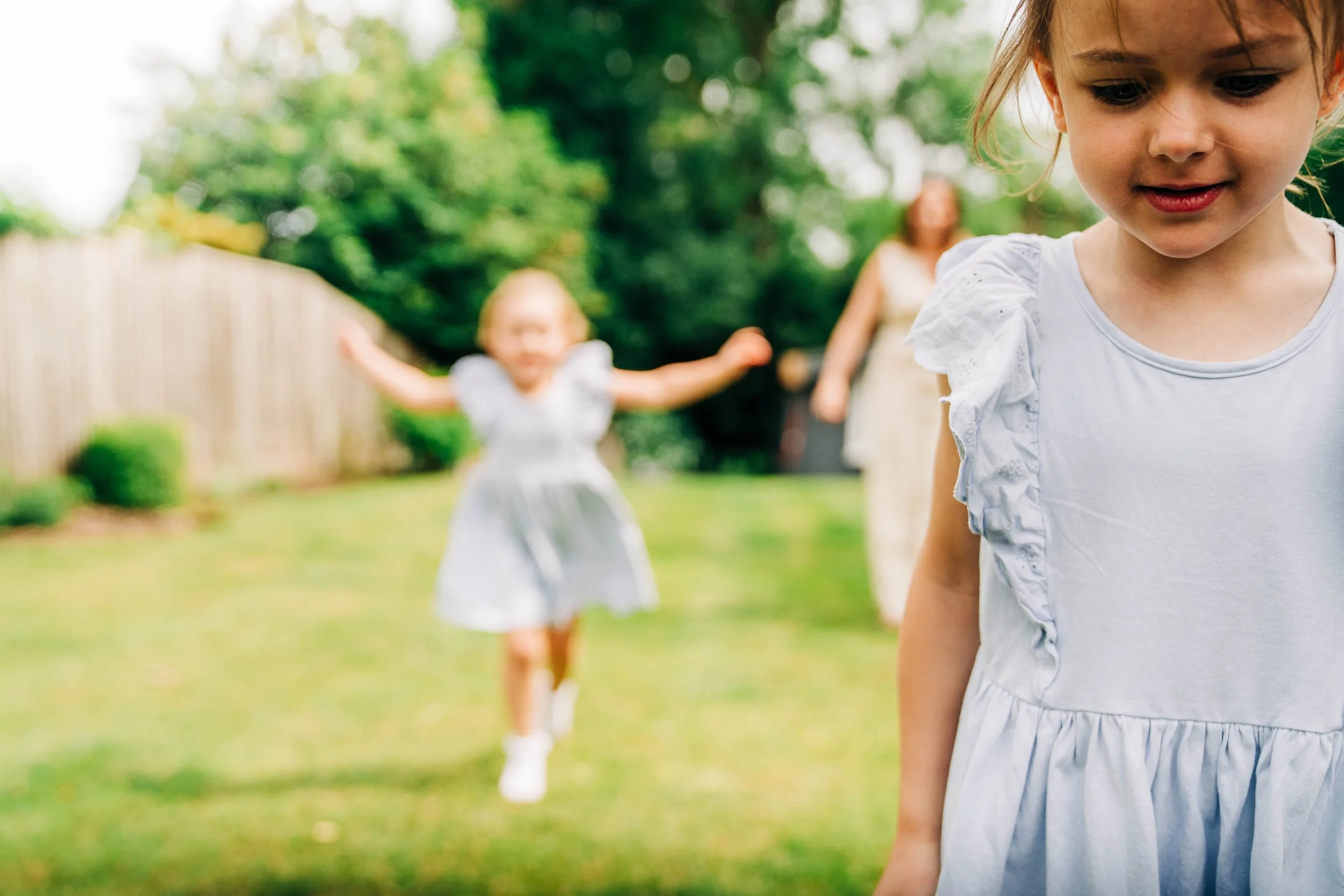 Two young girls in white dresses playing outdoors in a green backyard with a woman in the background. One girl is in the foreground looking down, and the other girl is blurred and running in the background.