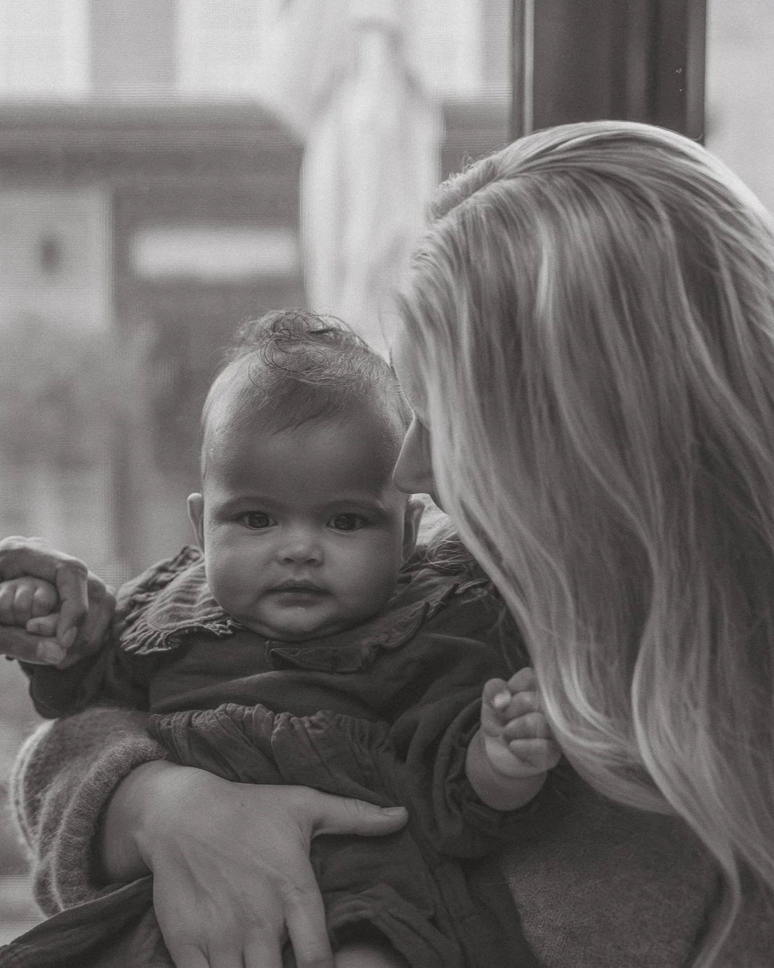 A woman with long hair holding a baby boy in a black and white photo. The woman appears to be leaning close to the baby, who is looking directly at the camera with a serious expression.