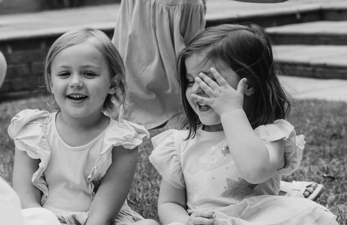 Two young girls sitting outdoors, laughing and smiling. One girl has short light hair and is looking at the camera, while the other girl has longer dark hair and is covering her face with her hand.
