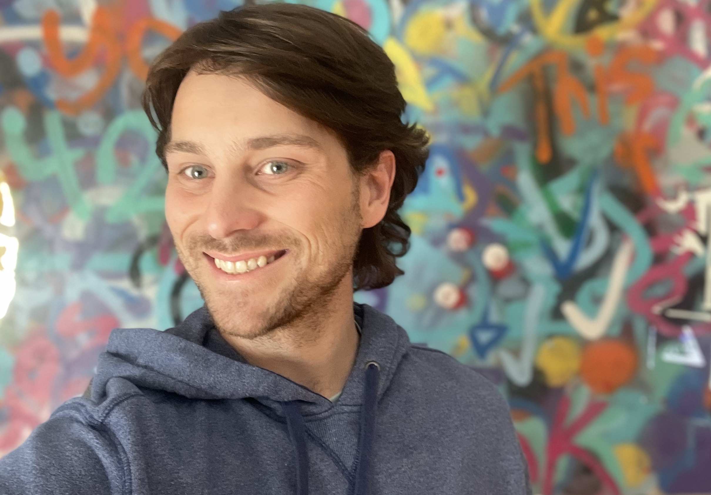 A smiling Kam with medium-length dark brown hair and light eyes is taking a selfie in front of an abstract, colorful self-painted graffiti wall.