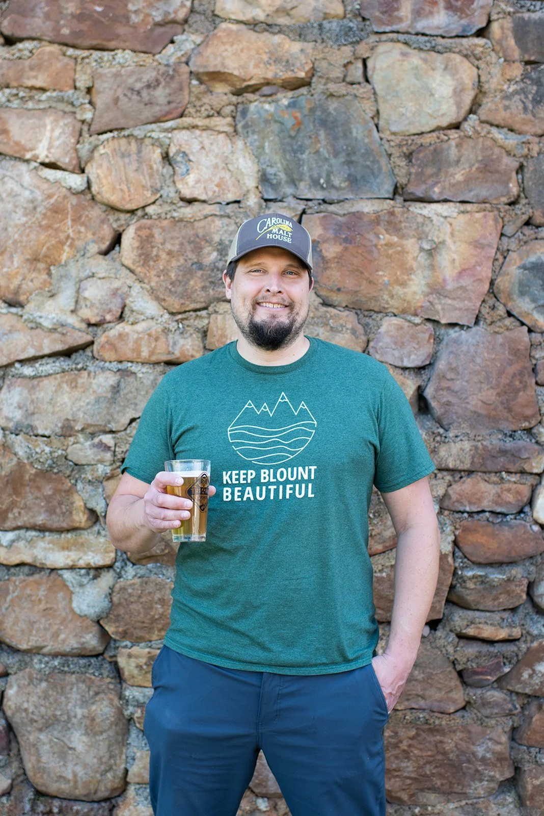 A man smiling and holding a glass of beer, standing in front of a stone wall, wearing a green T-shirt with mountain and water design and the text 'Keep Blount Beautiful', and a baseball cap with the logo 'Carolina Malt House'.