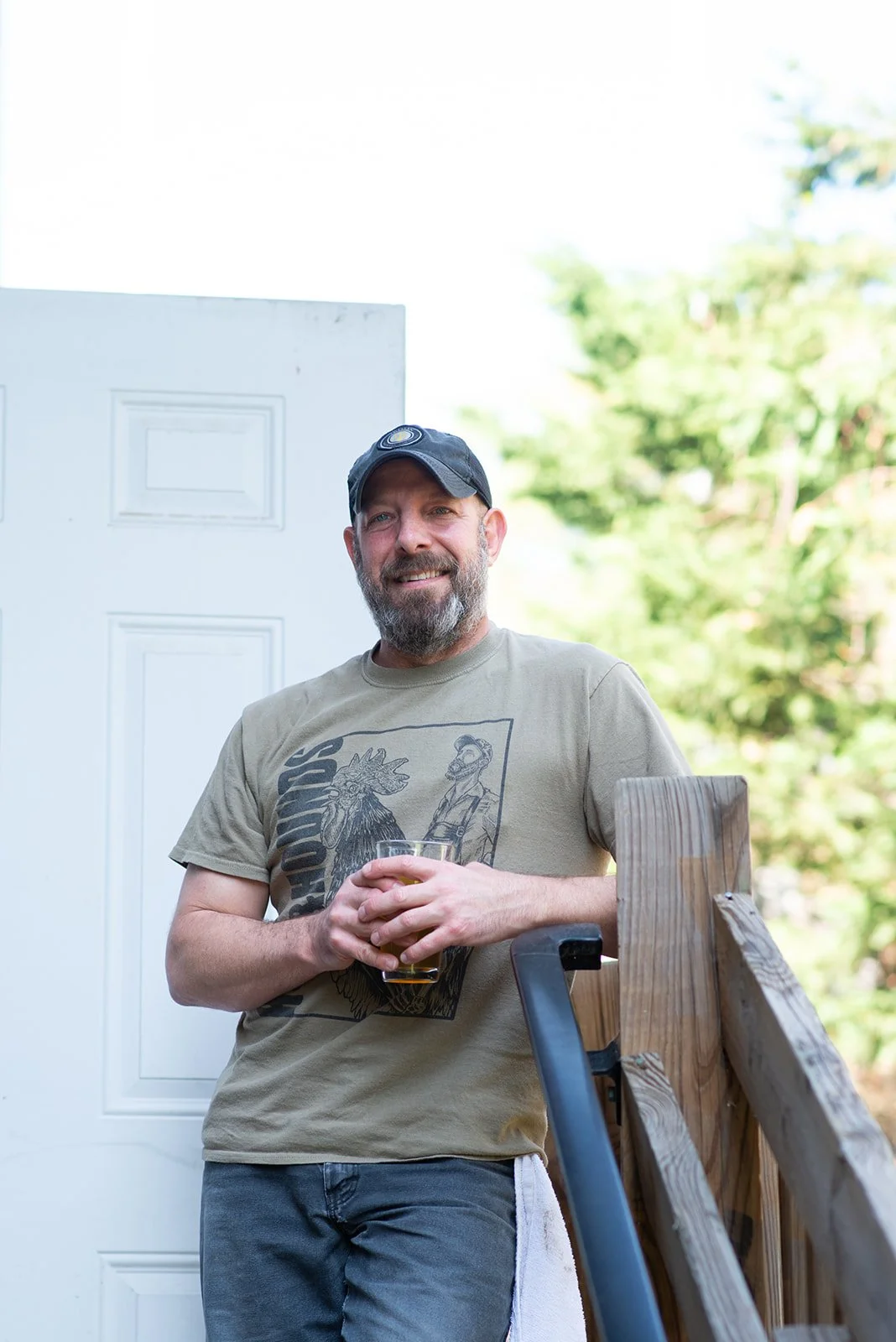 A man with a beard and a cap, smiling, standing outdoors near a wooden railing, holding a glass of beer, with green trees and a white door behind him.