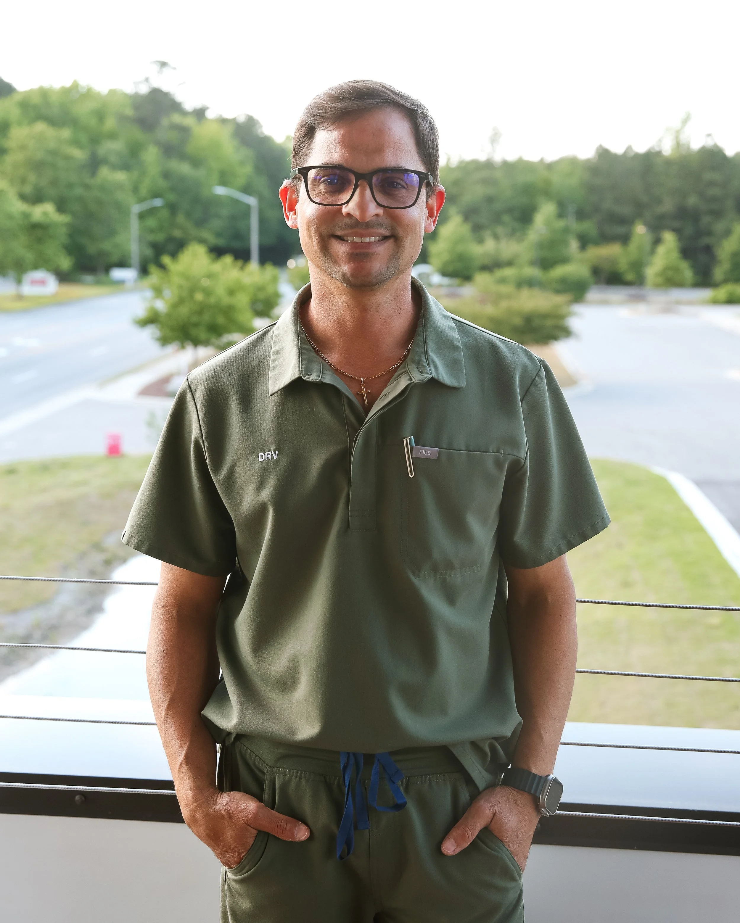 A man in a green uniform with glasses, standing outdoors with trees and a road in the background.