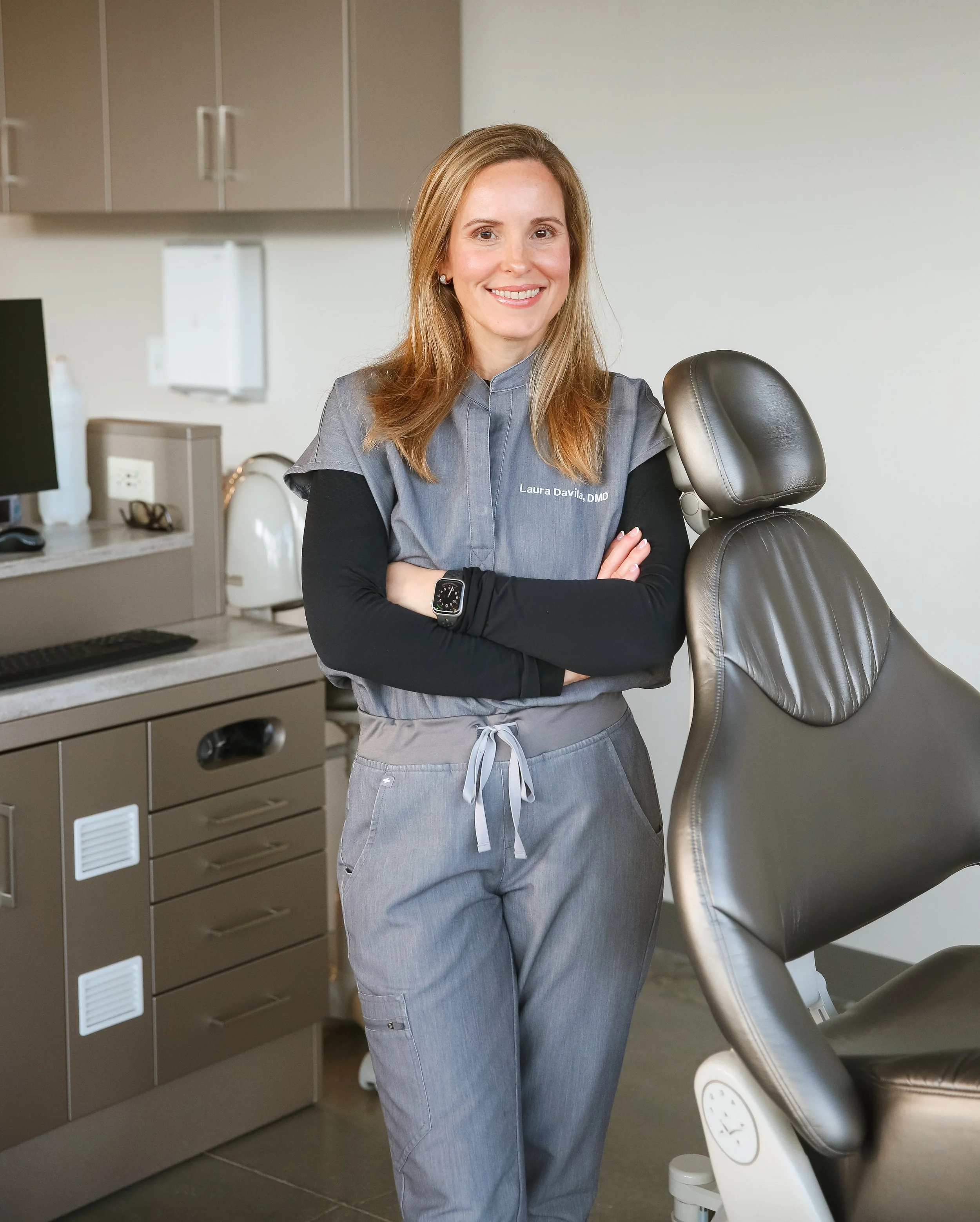 A female dentist in gray scrubs standing in a dental clinic, smiling with arms crossed, next to a dental chair.