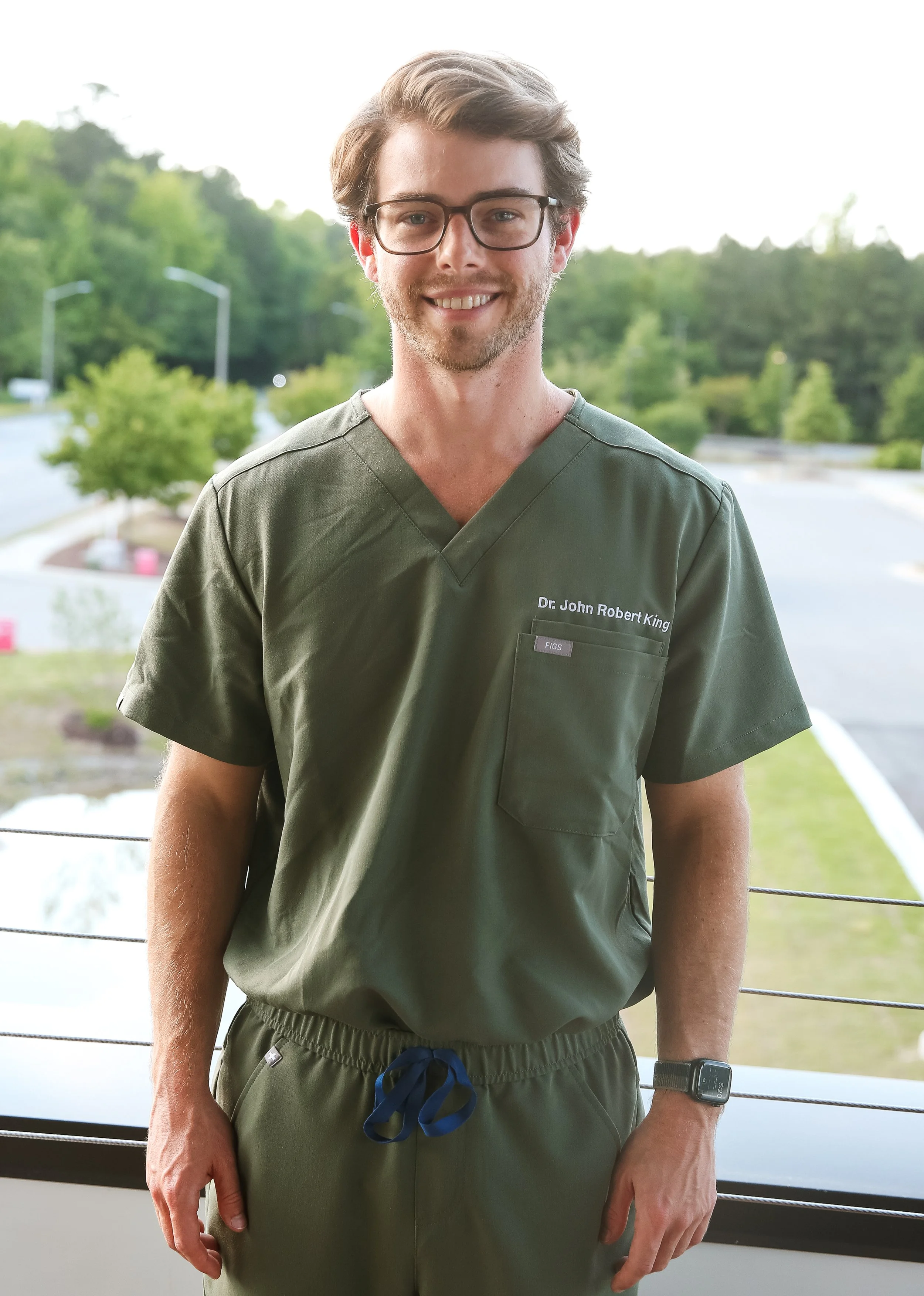 A man wearing glasses and green medical scrubs with a name tag that reads Dr. John Robert King, standing outdoors near a railing with trees and a parking lot in the background.