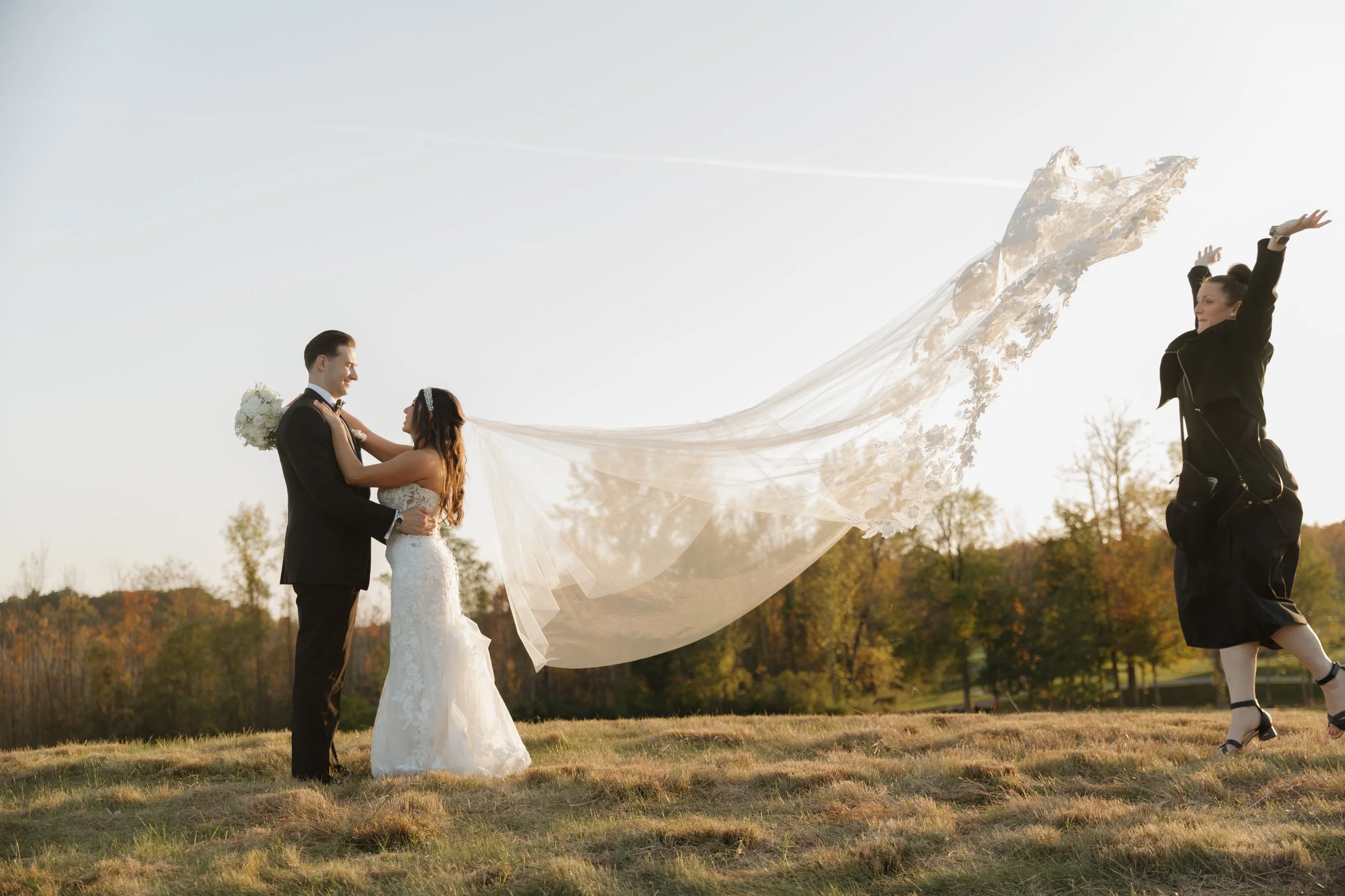 A bride and groom are standing outdoors in a field, facing each other, with a wedding veil flowing behind them. A photographer is capturing the moment. The trees in the background suggest it is autumn.