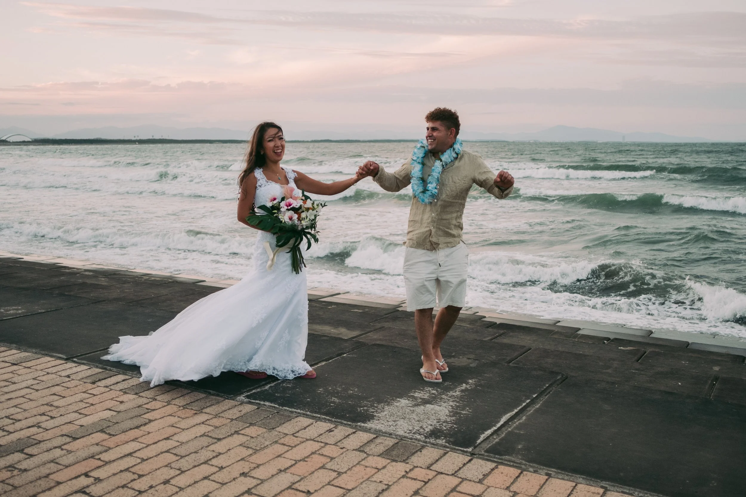 A couple celebrating by the beach, the woman in a wedding dress holding a bouquet, and the man in casual summer clothing with a blue lei, holding hands and smiling at each other.