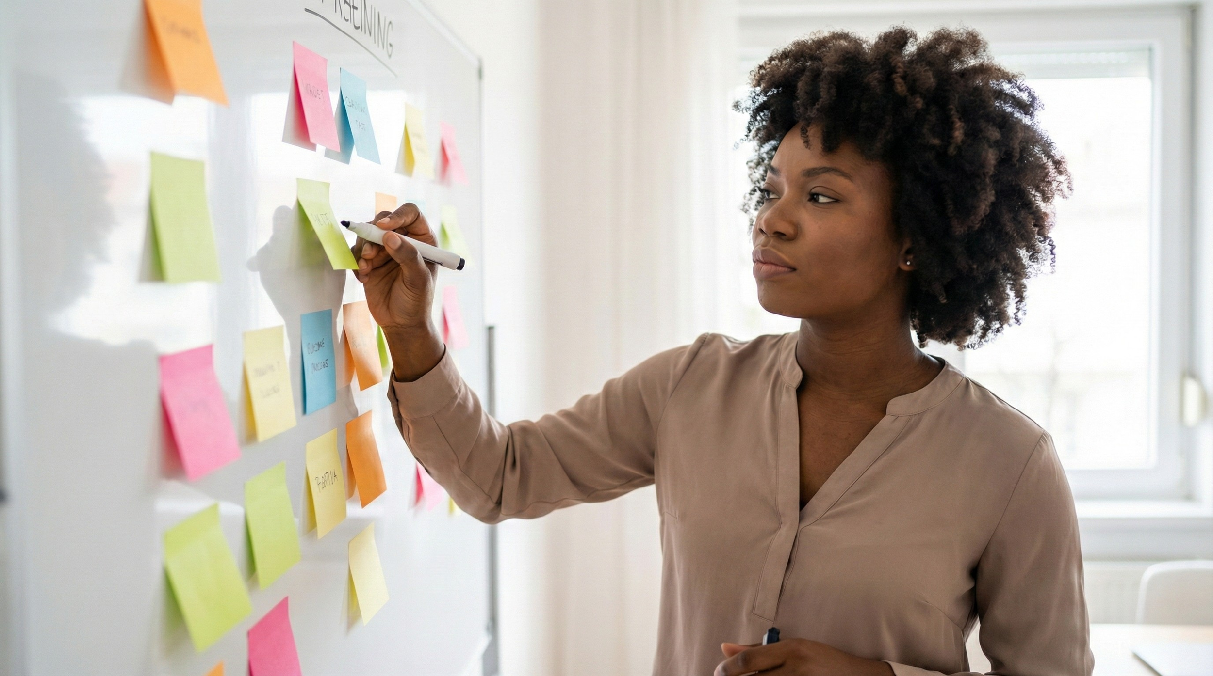 A professional Black woman using sticky notes on a whiteboard to organize her goals and systems, representing sustainable planning and alignment over simple busyness.