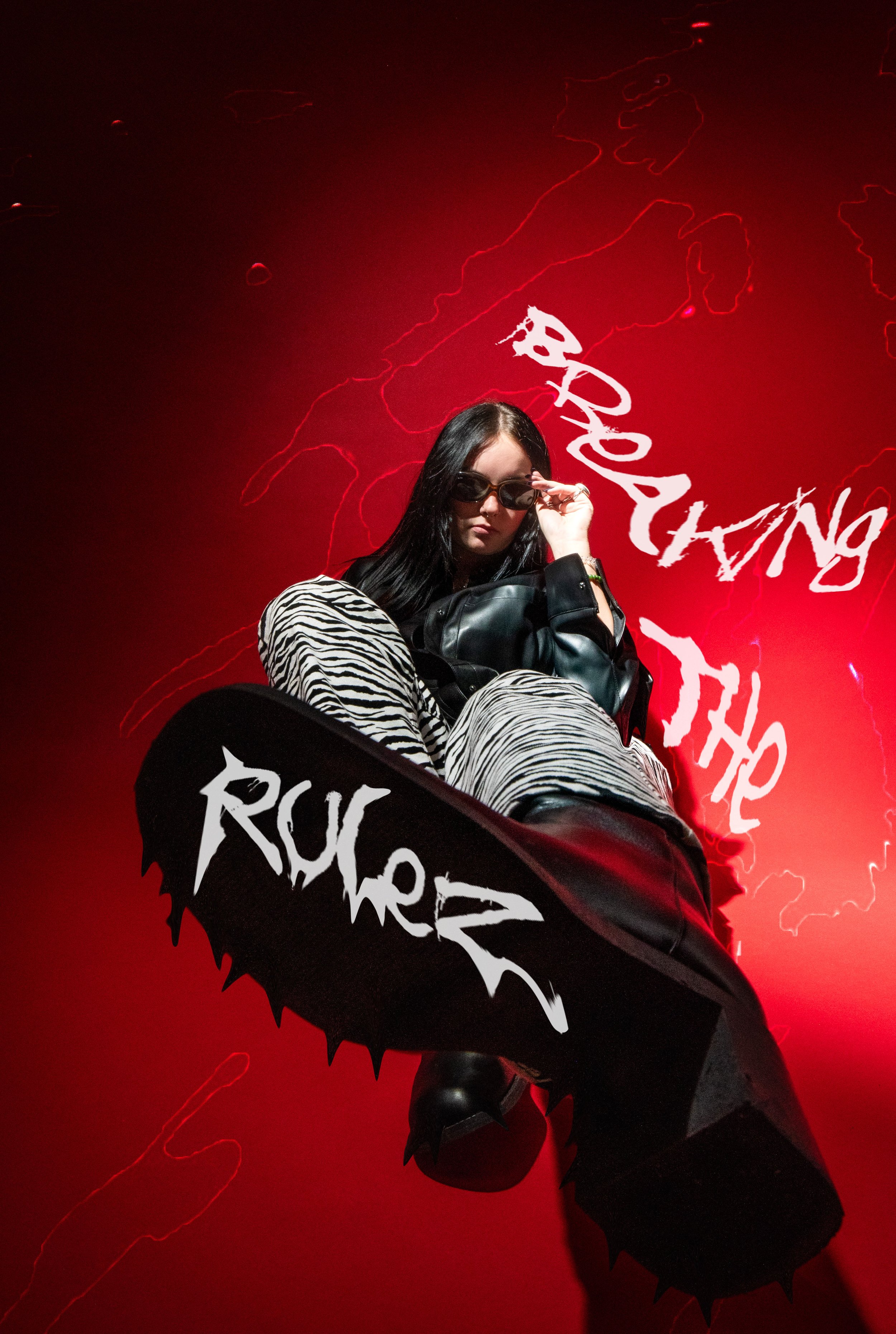 A person with long dark hair wearing sunglasses, zebra print pants, a black leather jacket, and black shoes, sitting with their foot extended toward the camera. The background is red with abstract lightning-like lines and the words 'Rukwei' and 'The'
