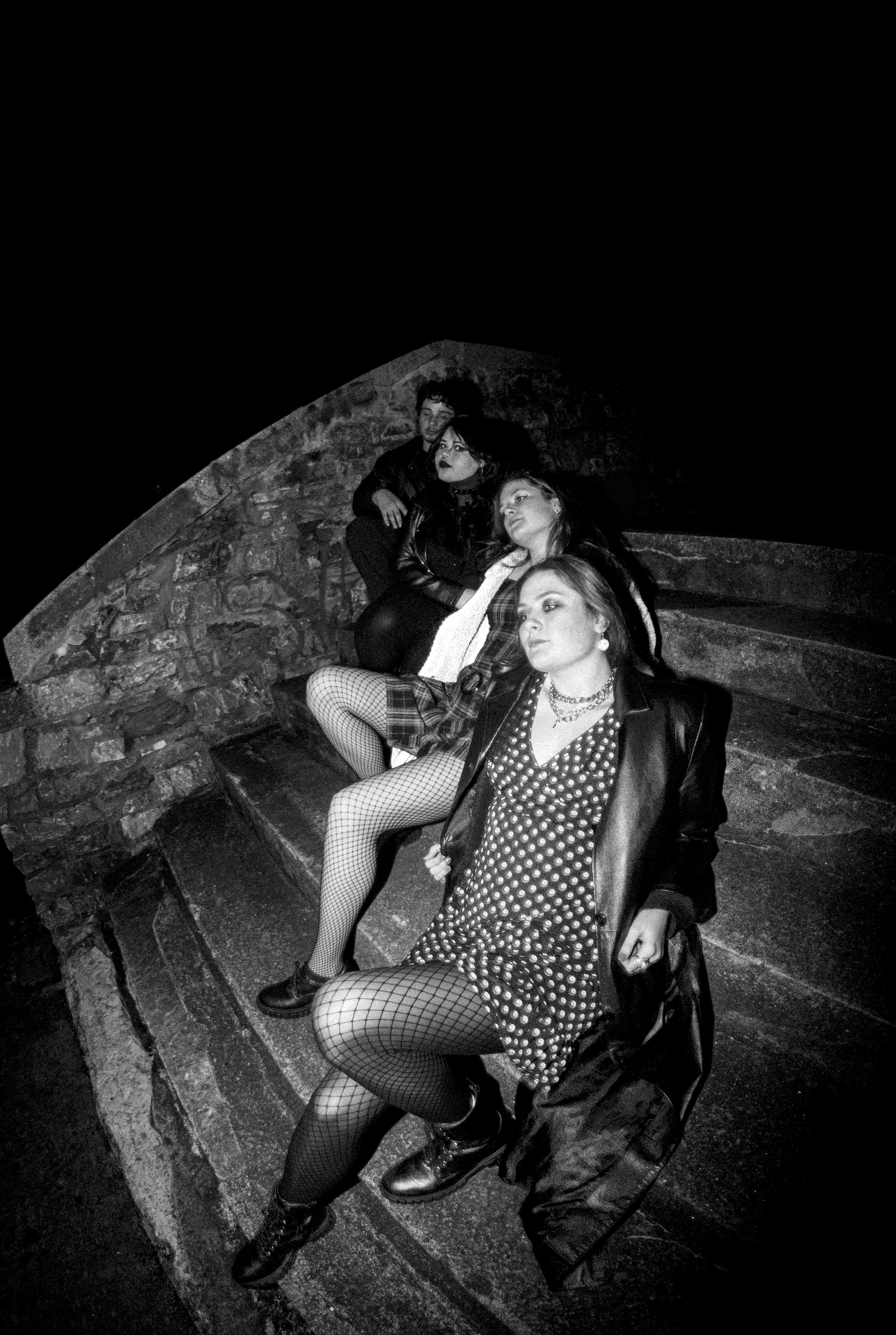 Black and white photo of four young women sitting on stairs under an arch, dressed in punk rock style with leather jackets, fishnet stockings, and jewelry.