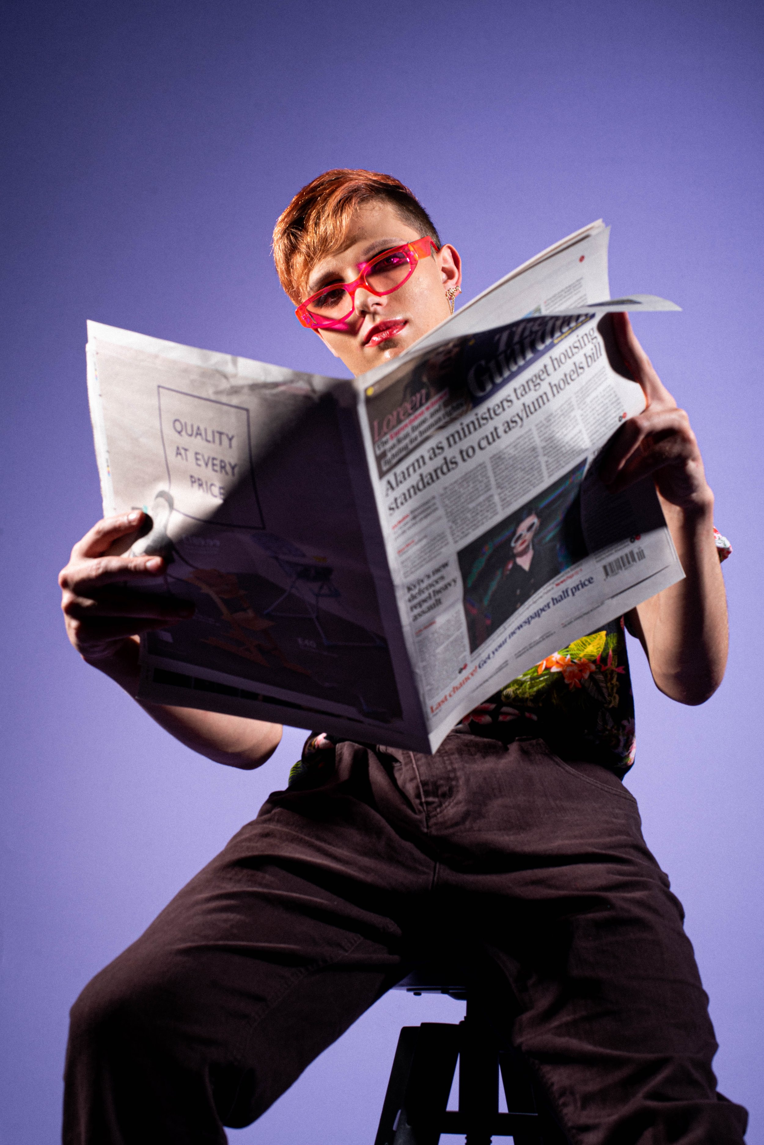 Person with short hair, pink glasses, and colorful clothing sitting on a stool, reading a newspaper against a purple background.