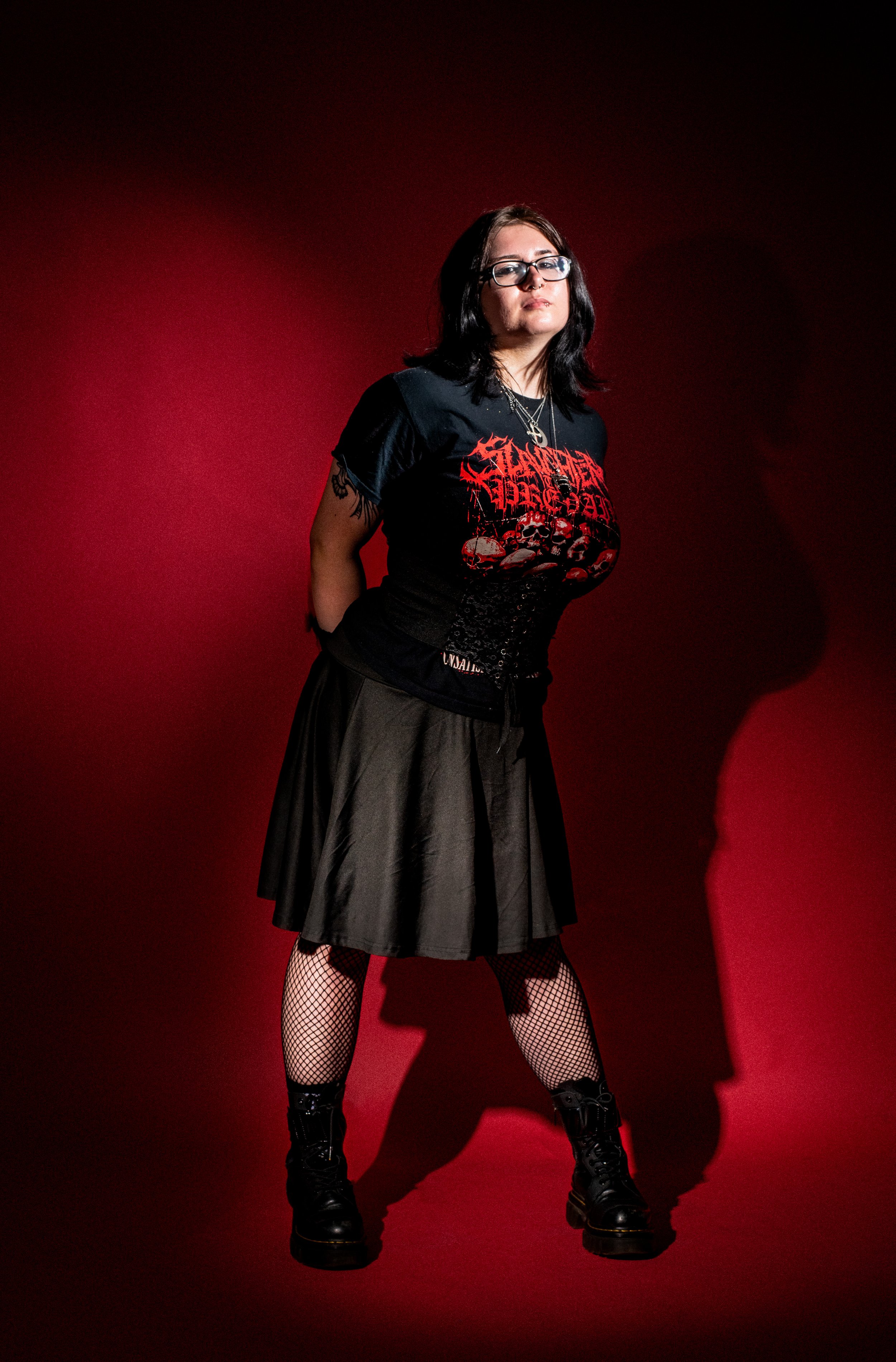 A woman with black hair and glasses standing against a red background, wearing a black graphic t-shirt, black skirt, fishnet stockings, and black combat boots.