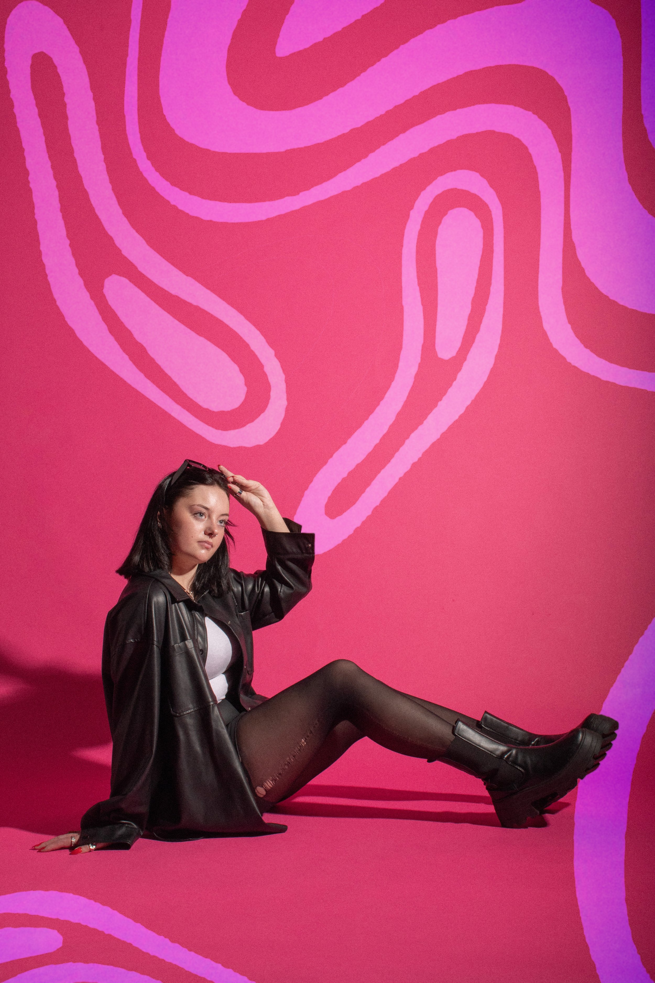 A young woman with dark hair and light skin sitting on a pink floor, wearing a black leather jacket, black tights, black boots, and sunglasses on her head, with a pink background featuring abstract purple and pink lines.