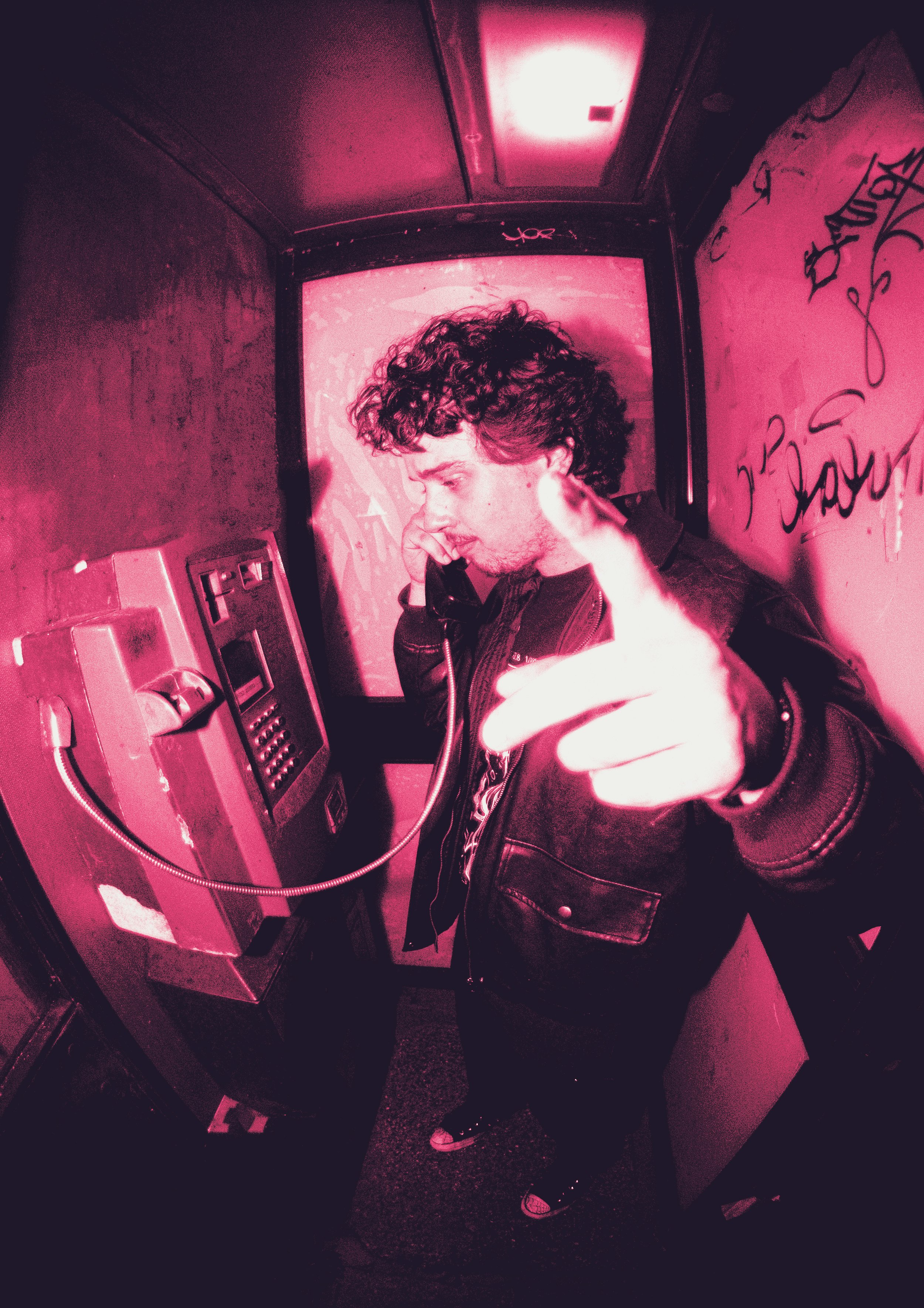 A young man with curly hair answering a payphone in a small, graffiti-covered phone booth, pointing at the camera.