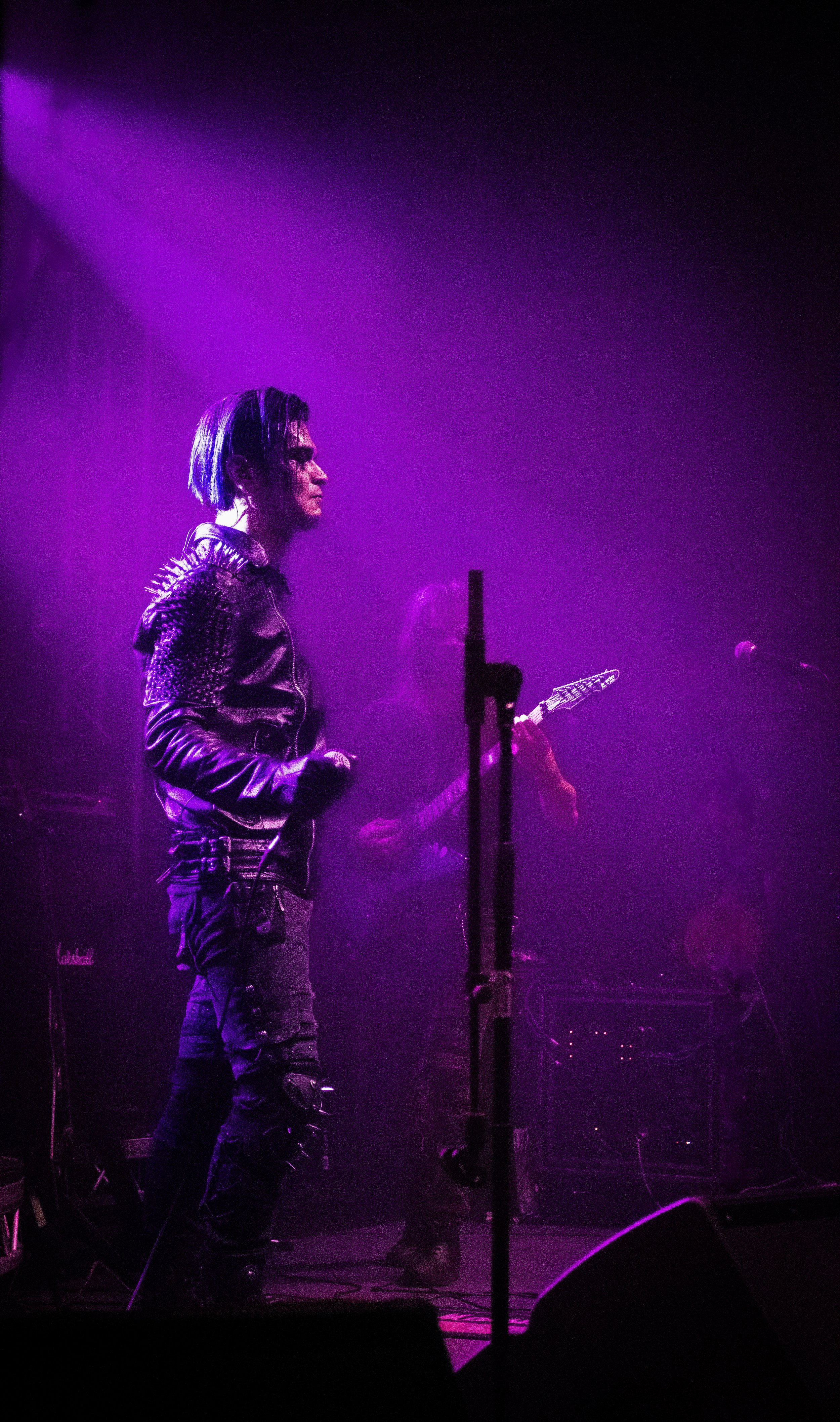 A musician with short hair and a leather jacket performing on stage with purple lighting, holding a microphone, and another person in the background playing an electric guitar.