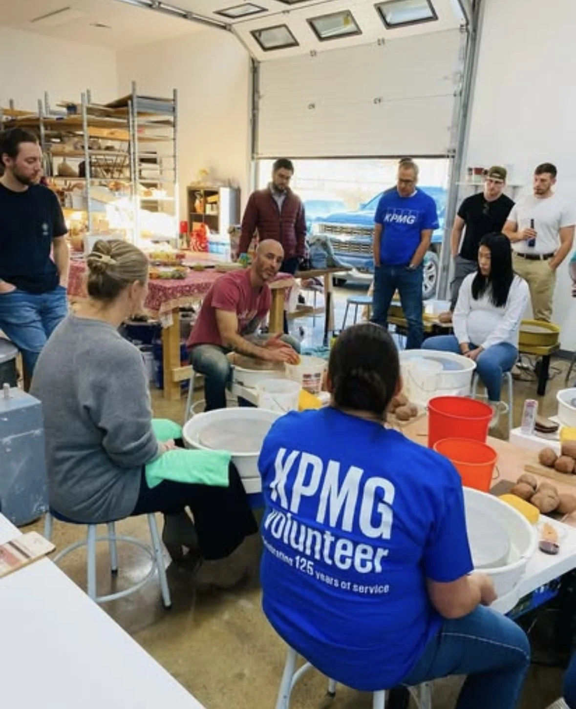 A group of people gathered around a table in a workshop, with some sitting and some standing, while one person demonstrates handling potatoes near a pottery wheel.