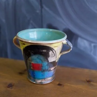 A ceramic vessel with a rounded body and vertical handles, unglazed with a matte brown finish, sitting on a blue spinning pottery wheel in a workshop.