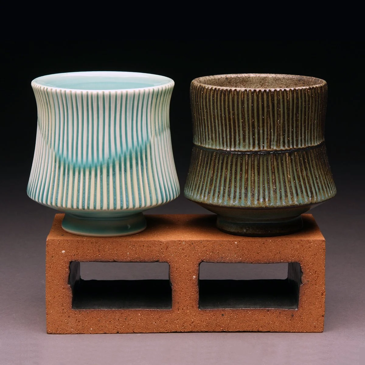 Two ceramic cups with vertical ribbed patterns, one in light green and white, the other in dark brown and green, are placed on a brick stand against a black background.