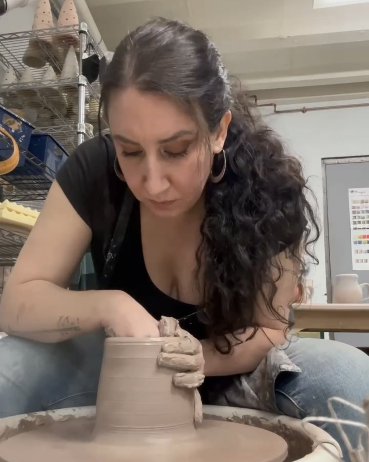 A person engaged in pottery, shaping clay on a pottery wheel, focused on the task. Shelves with pottery items in the background.