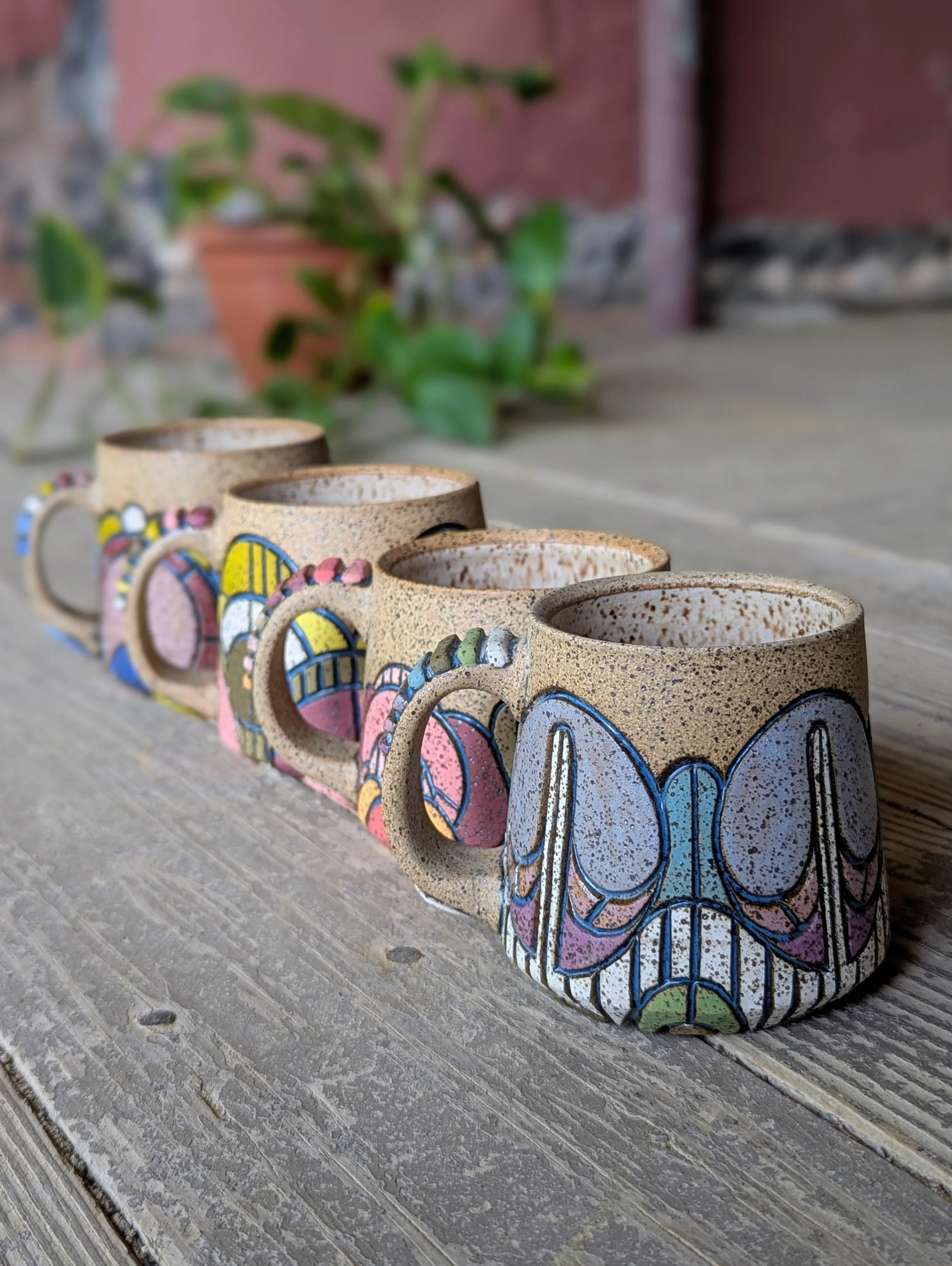 Four handcrafted ceramic mugs with colorful, abstract patterns are arranged in a row on a weathered wooden surface outdoors, with a potted plant and stone wall in the background.