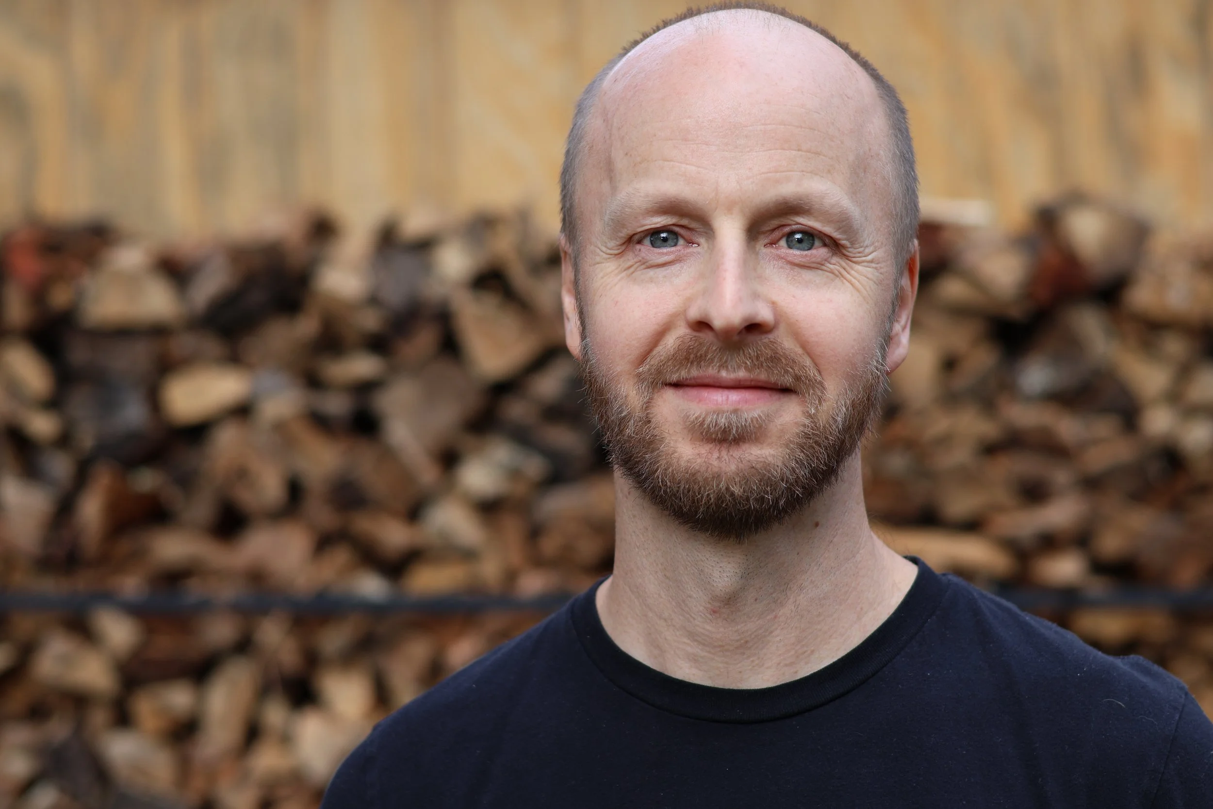 Close-up portrait of a man with a beard and blue eyes smiling outdoors with a background of stacked firewood.