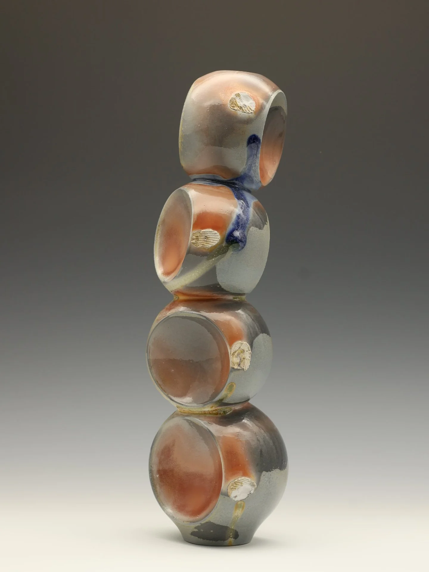 Stacked abstract ceramic sculptures with a glossy finish in shades of beige, orange, gray, and blue on a reflective surface against a gray background.