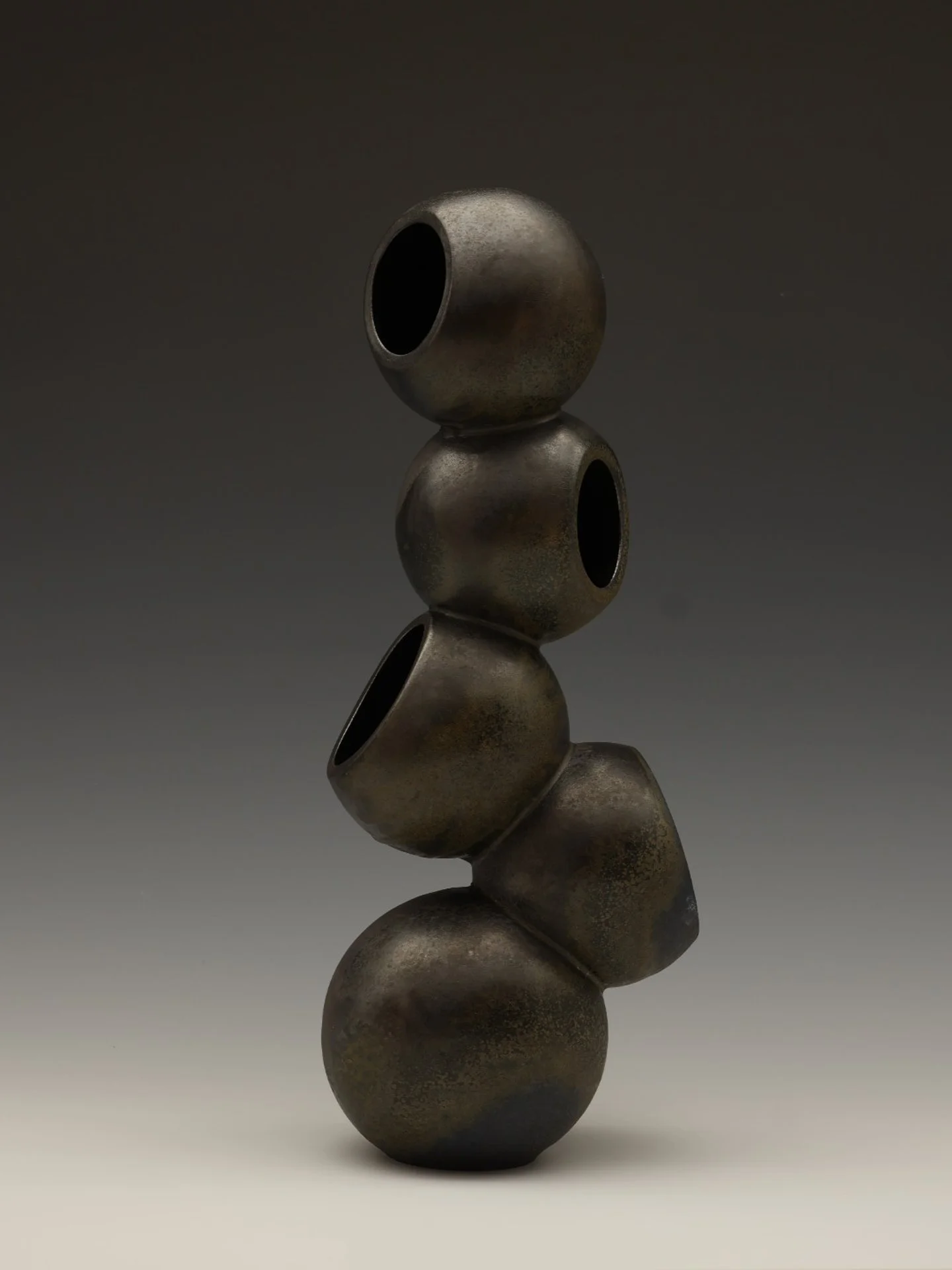 A vertical stack of five rounded, metallic, hollow spheres with a weathered, dark surface, arranged against a dark background.