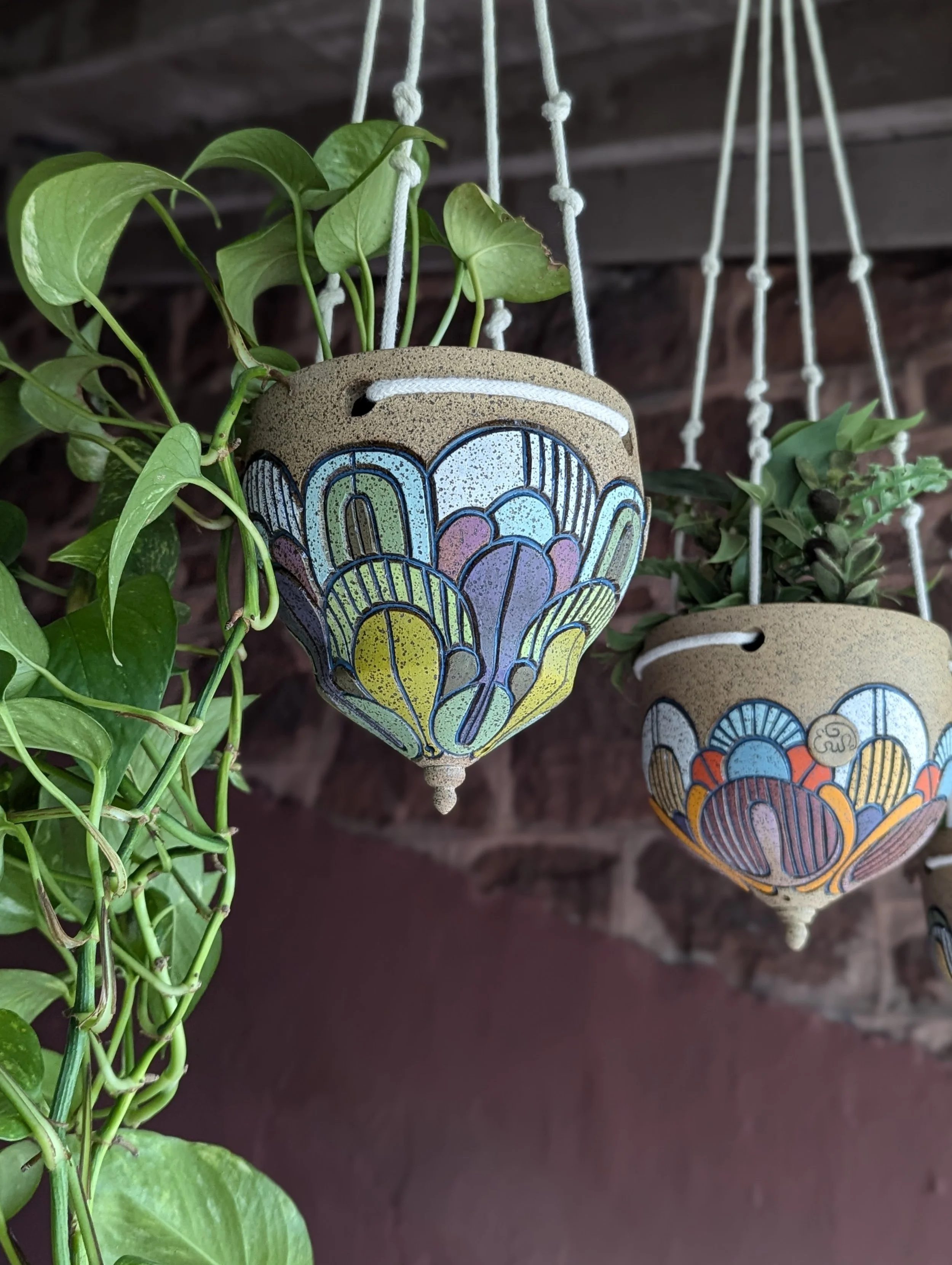 Colorful ceramic hanging flower pots with painted floral designs, hanging next to green plants against a brick wall.